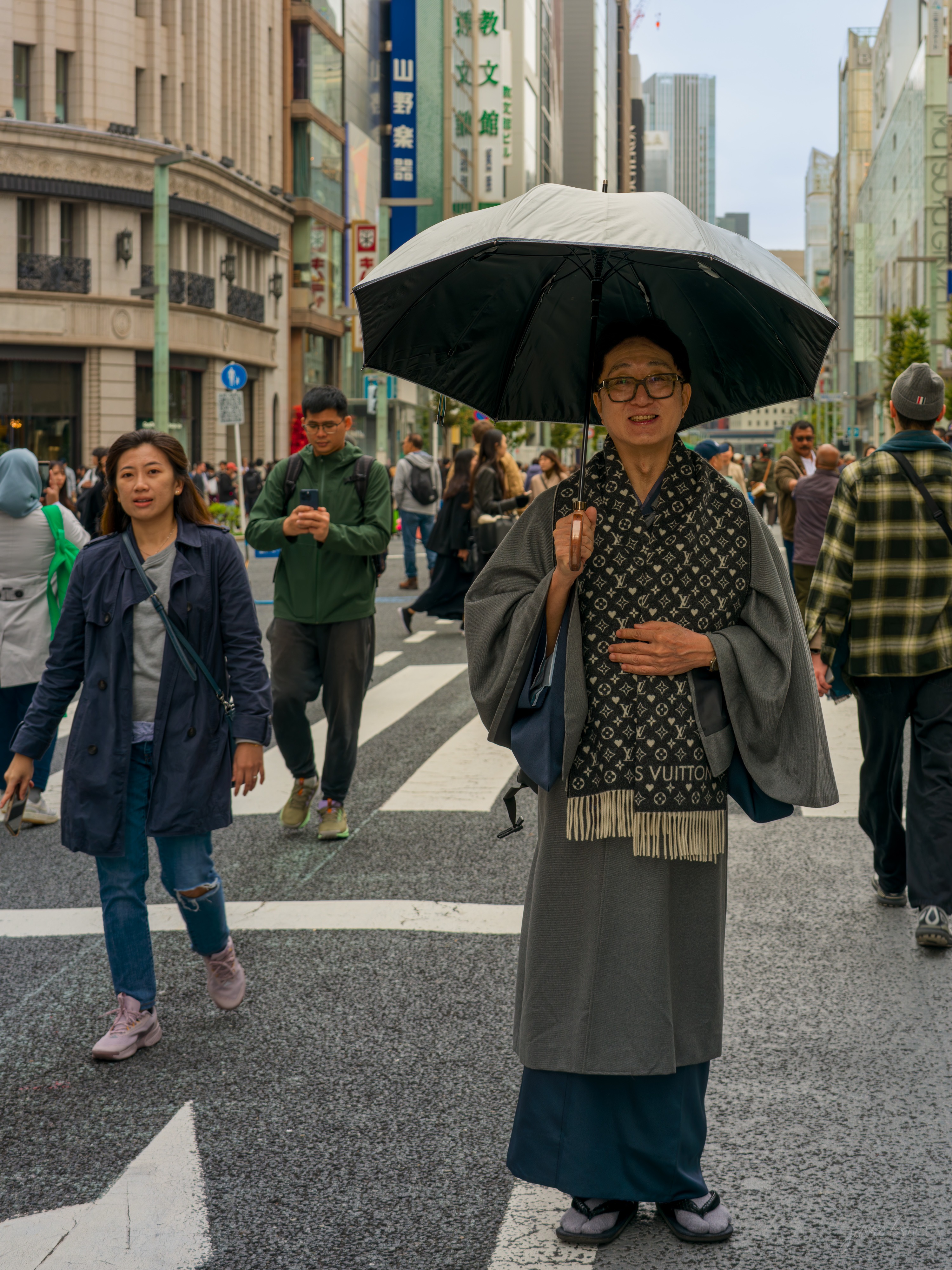 A man stands in the middle of a wide pedestrian crossing on a busy shopping street, holding a clear-topped umbrella overhead. He wears a dark kimono and haori printed with a repeating monogram and a sash reading Louis Vuitton, one hand raised, looking toward the camera. Shoppers cross around him.
