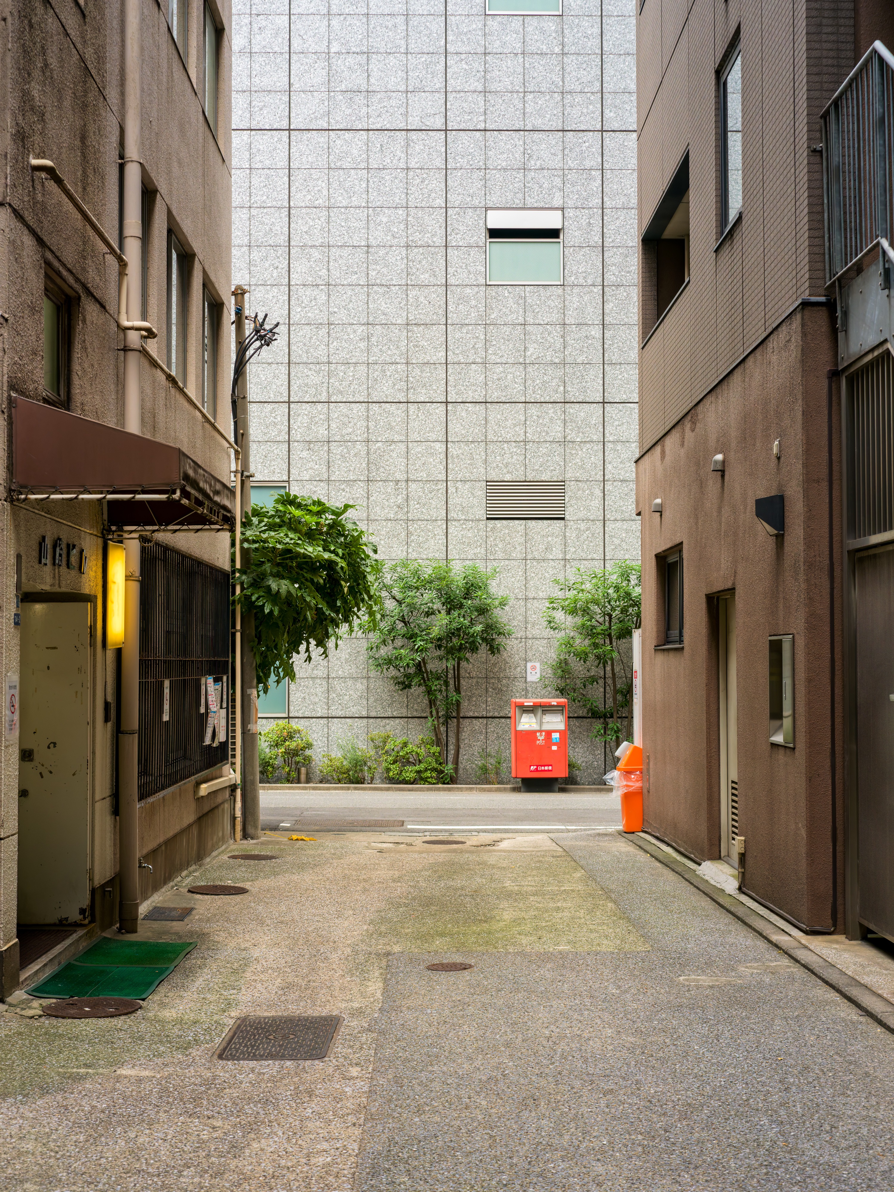 A quiet paved backstreet between buildings, ending at a large blank tiled wall. A red Japanese postbox stands against the wall beside a small tree, with an orange traffic cone next to it.