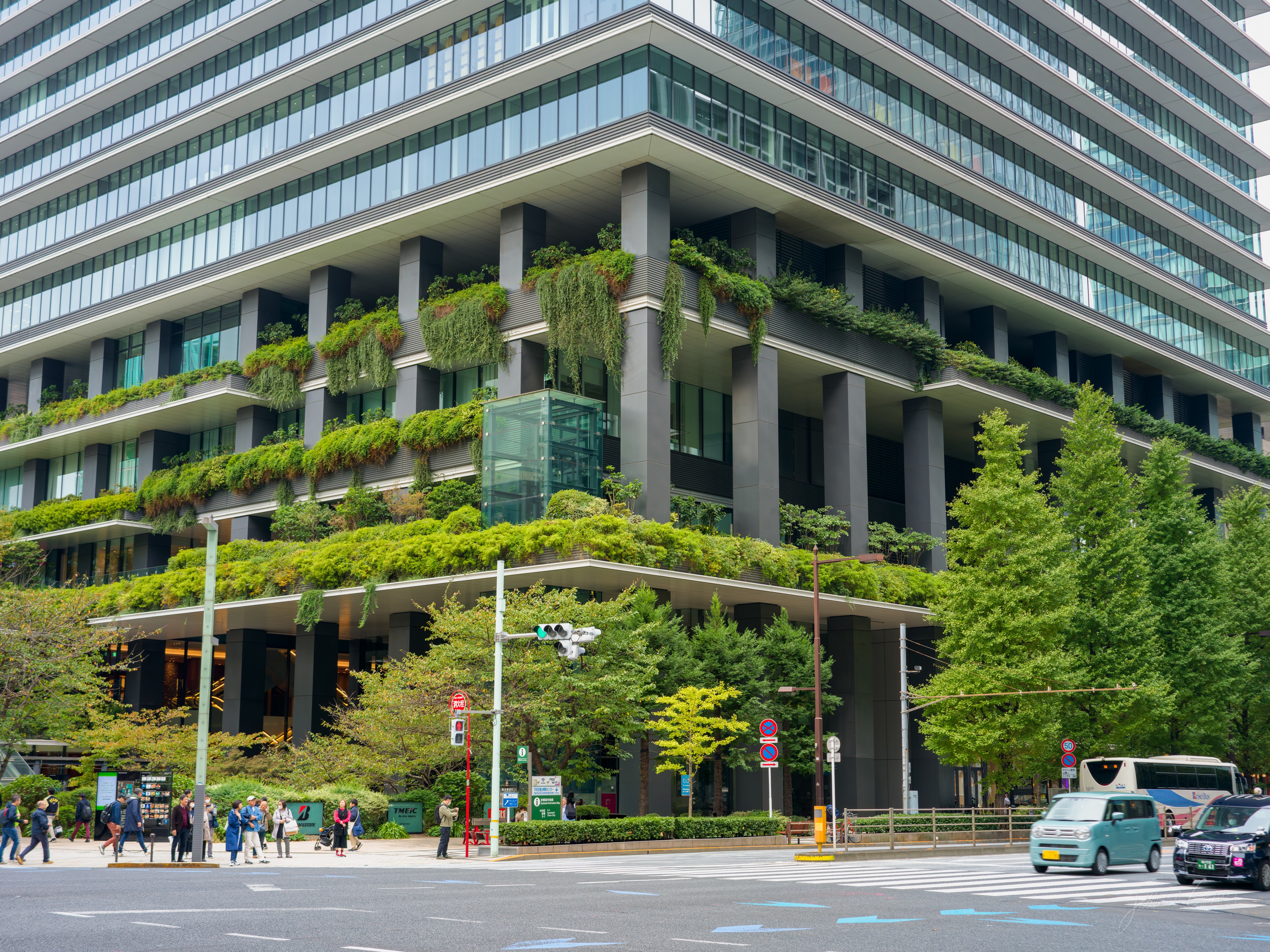 A large office building on a street corner with planted greenery cascading from the edge of every floor's terrace. People wait at the crosswalk in front and a small van and a taxi pass on the road.