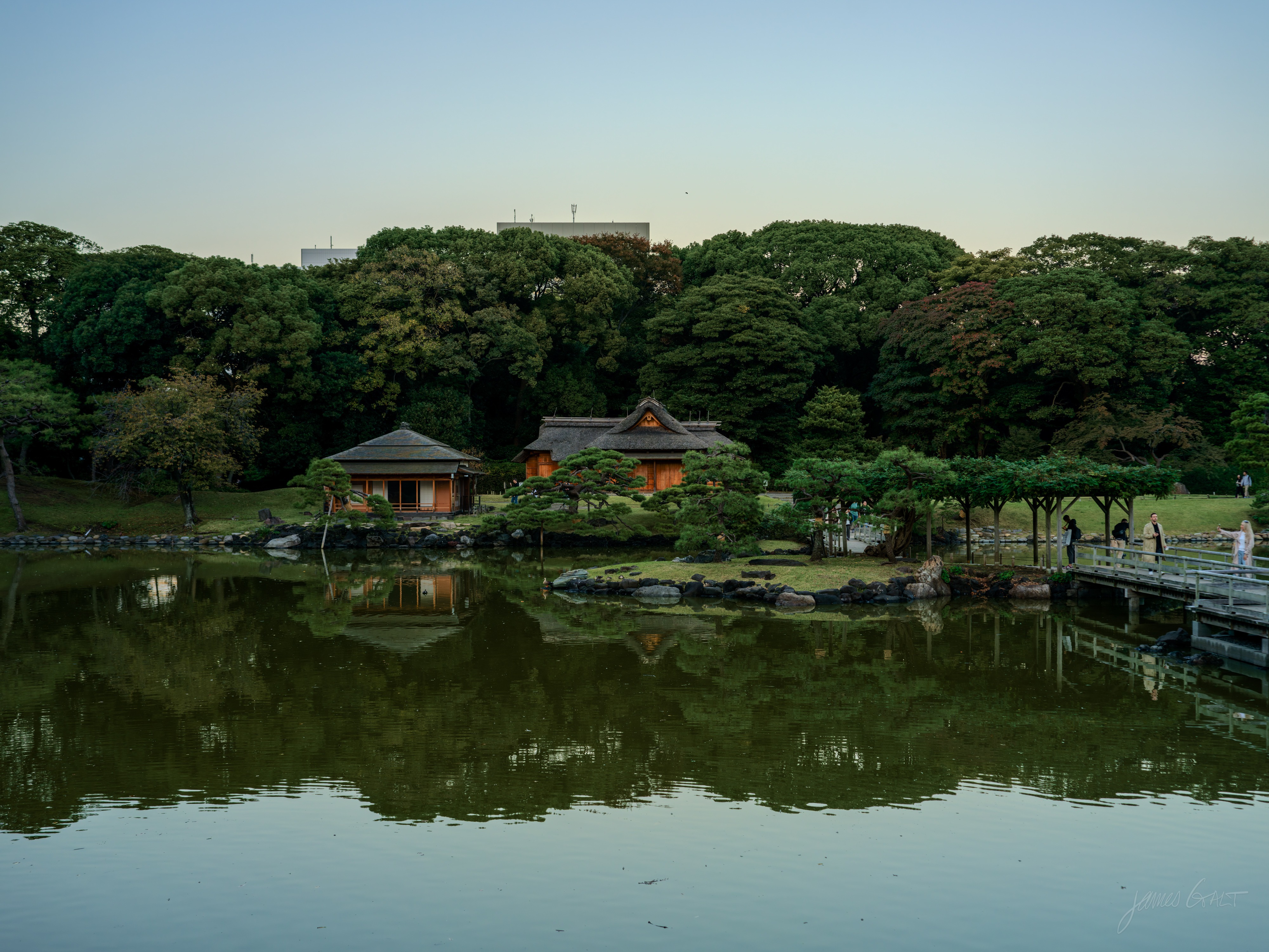 A calm pond at dusk reflecting a wooded far bank, where two low wooden teahouses, one thatched, sit at the water's edge among pines. A wooden trellis walkway runs along the right with a few people on it, and a small jetty extends into the water.