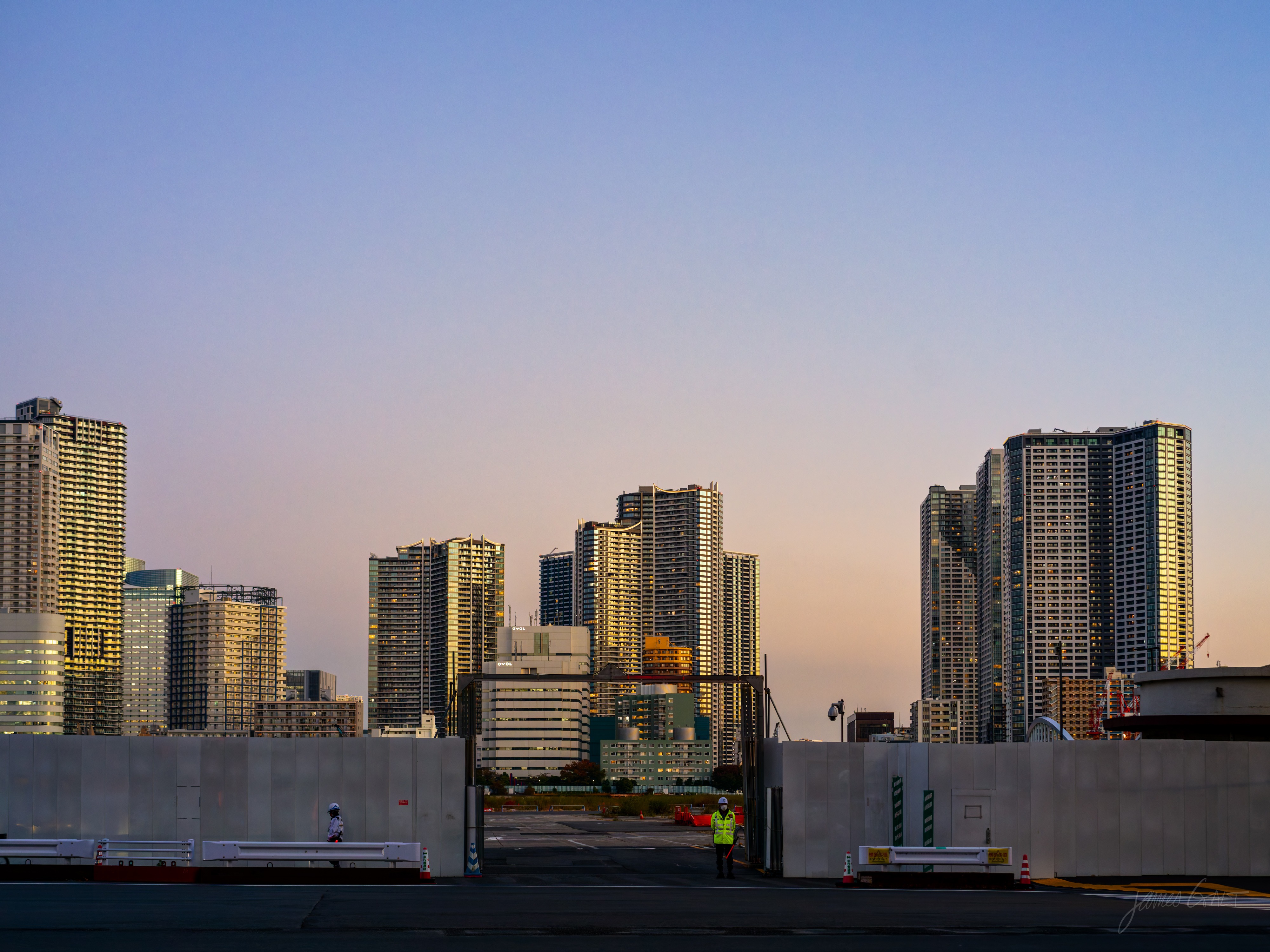 A dusk skyline of tall apartment towers catching the last warm sunlight at their tops against a graded pink-to-blue sky. In front, concrete hoarding walls enclose an empty paved lot where a lone worker in a hi-vis vest stands near traffic cones.