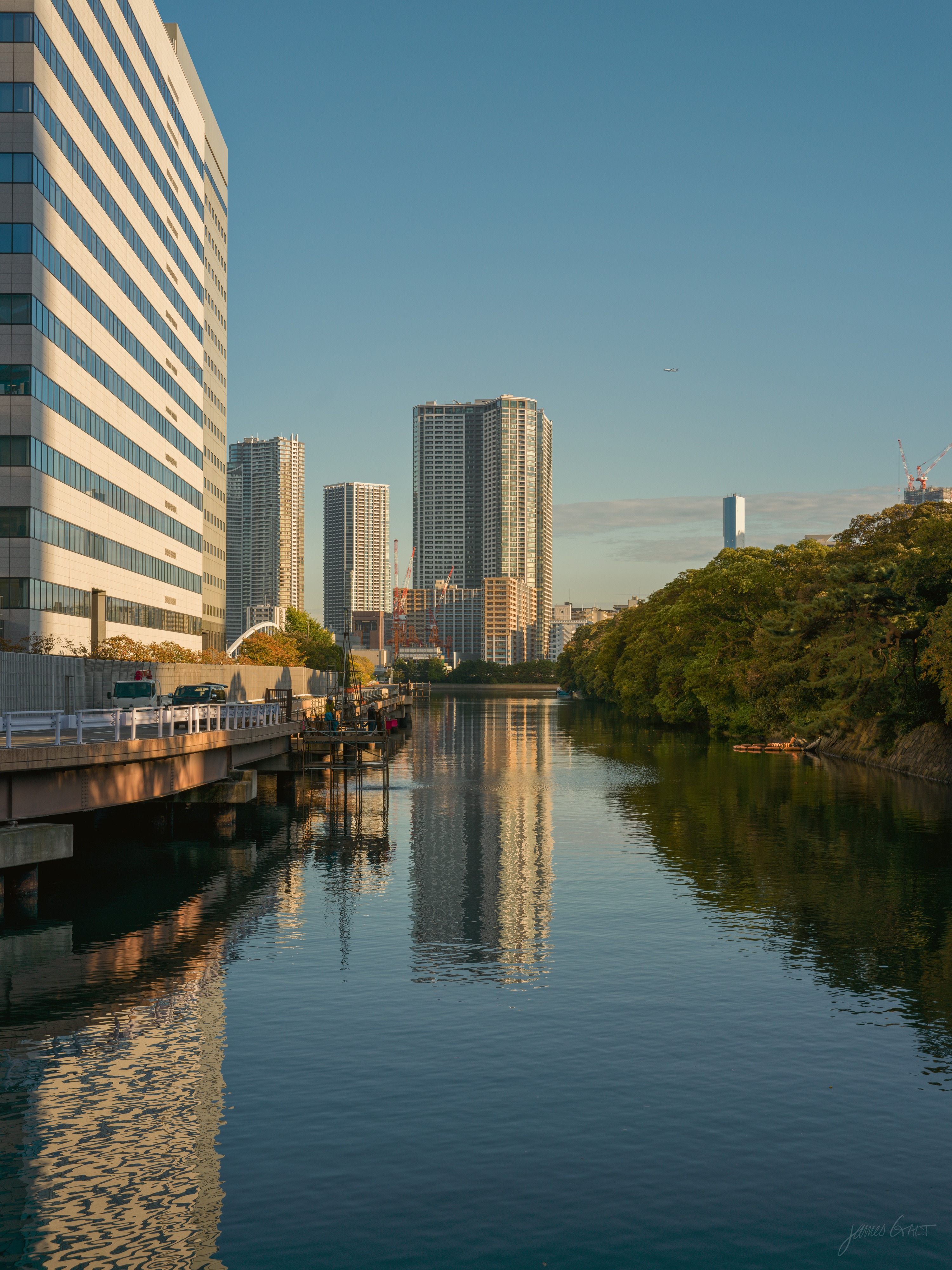 A still canal in golden late light, running between a long pale office building on the left and a wooded green bank on the right, with cars on an embankment. A cluster of tall apartment towers stands at the far end, reflected down the length of the water under a clear sky.