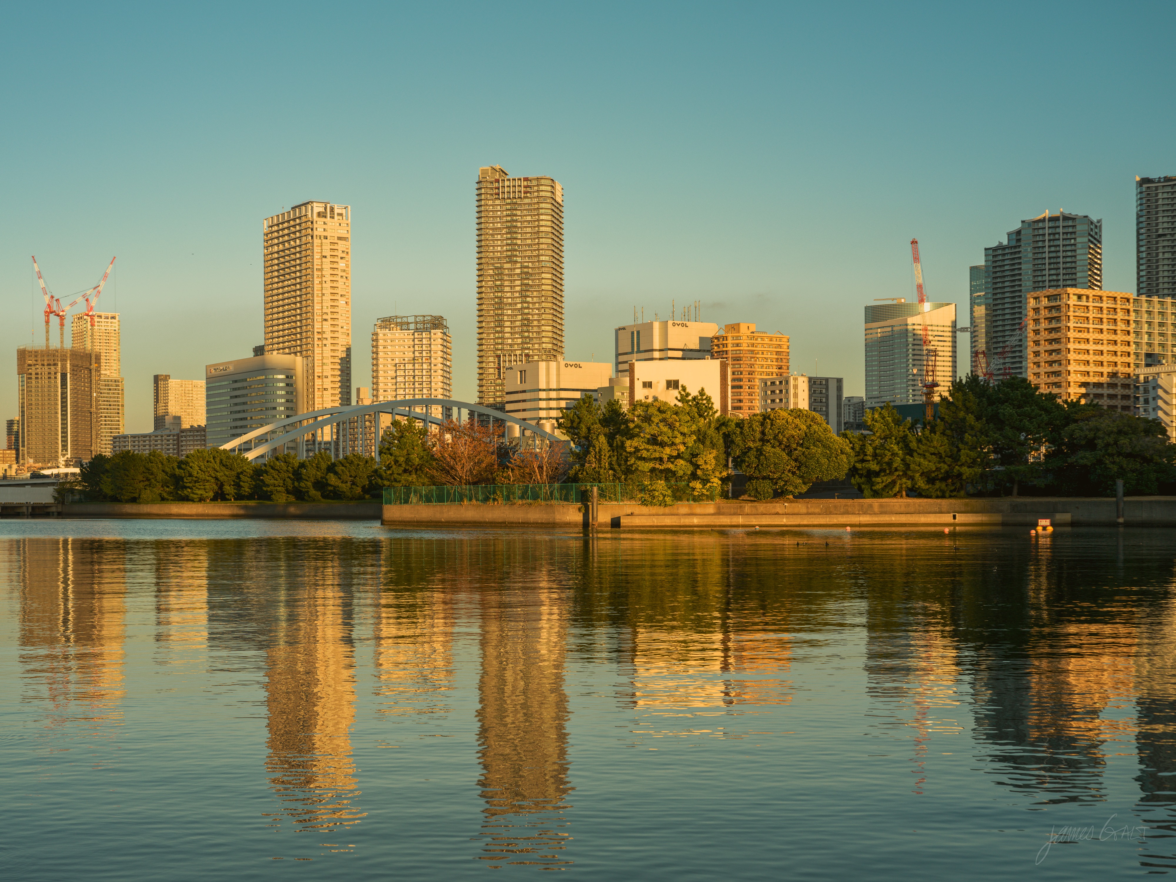A wide stretch of calm water at golden hour, with a city skyline of apartment and office towers and an arched bridge along the far shore, all lit warm and mirrored in the water below a pale sky.
