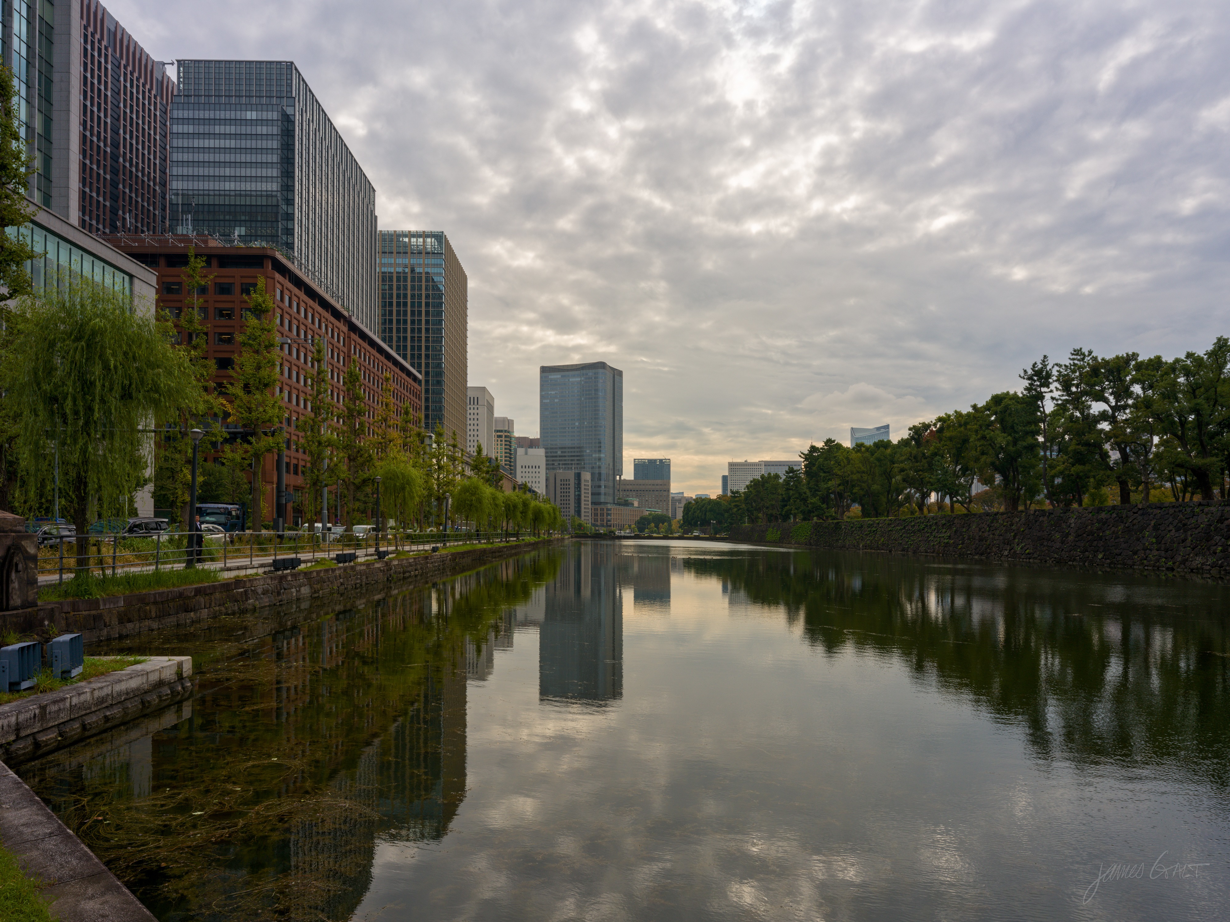 A wide moat reflecting an overcast sky, with the Imperial Palace's stone embankment and trees along the right bank and a row of office buildings, including a red-brick block and glass towers, along the left. Willows and a tree-lined path run beside the water.