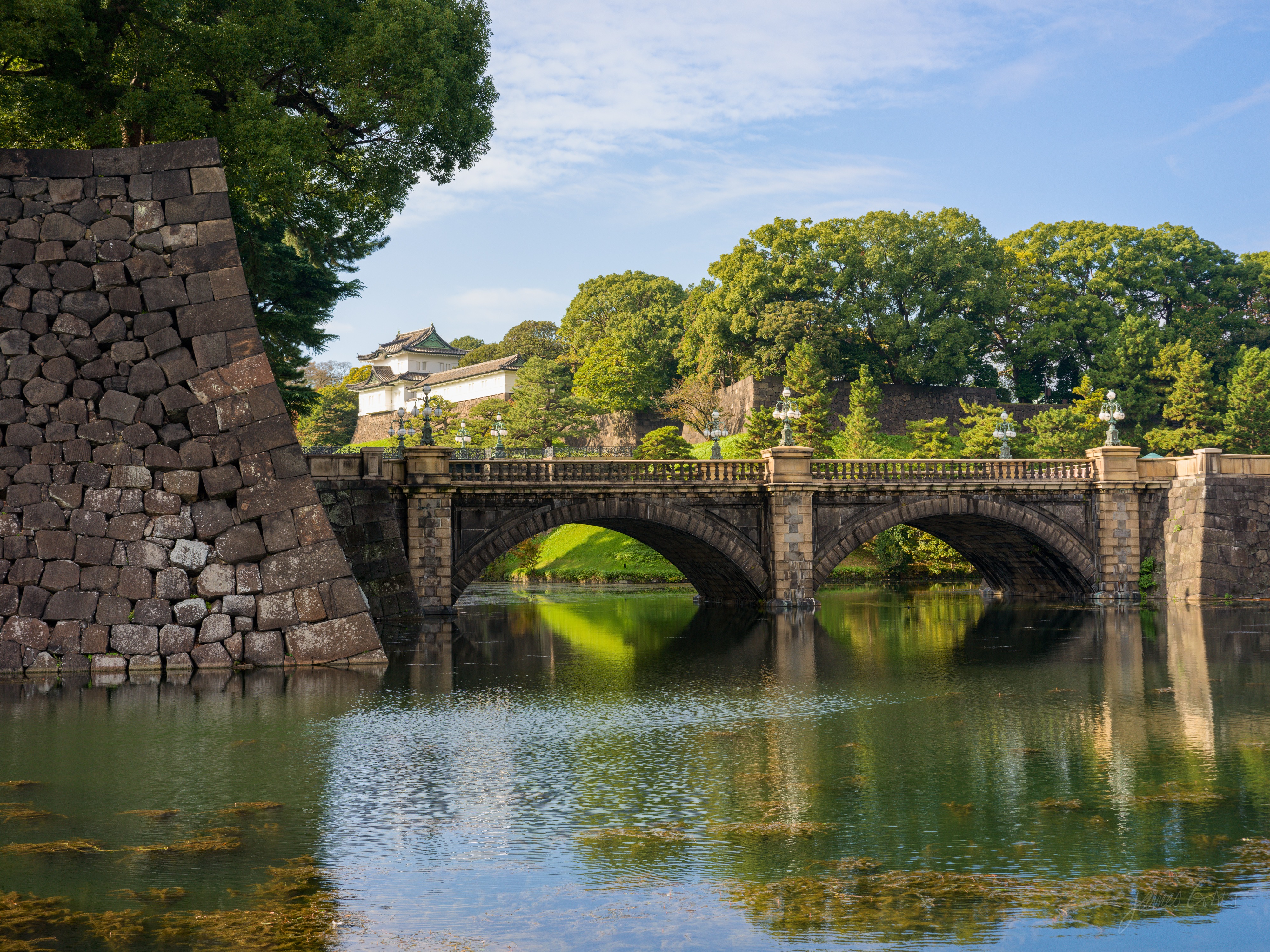 The Nijubashi stone bridge arching over the Imperial Palace moat, its two arches reflected in the water, with a sloping stone rampart on the left and a white-walled turret among trees behind, under a clear blue sky.