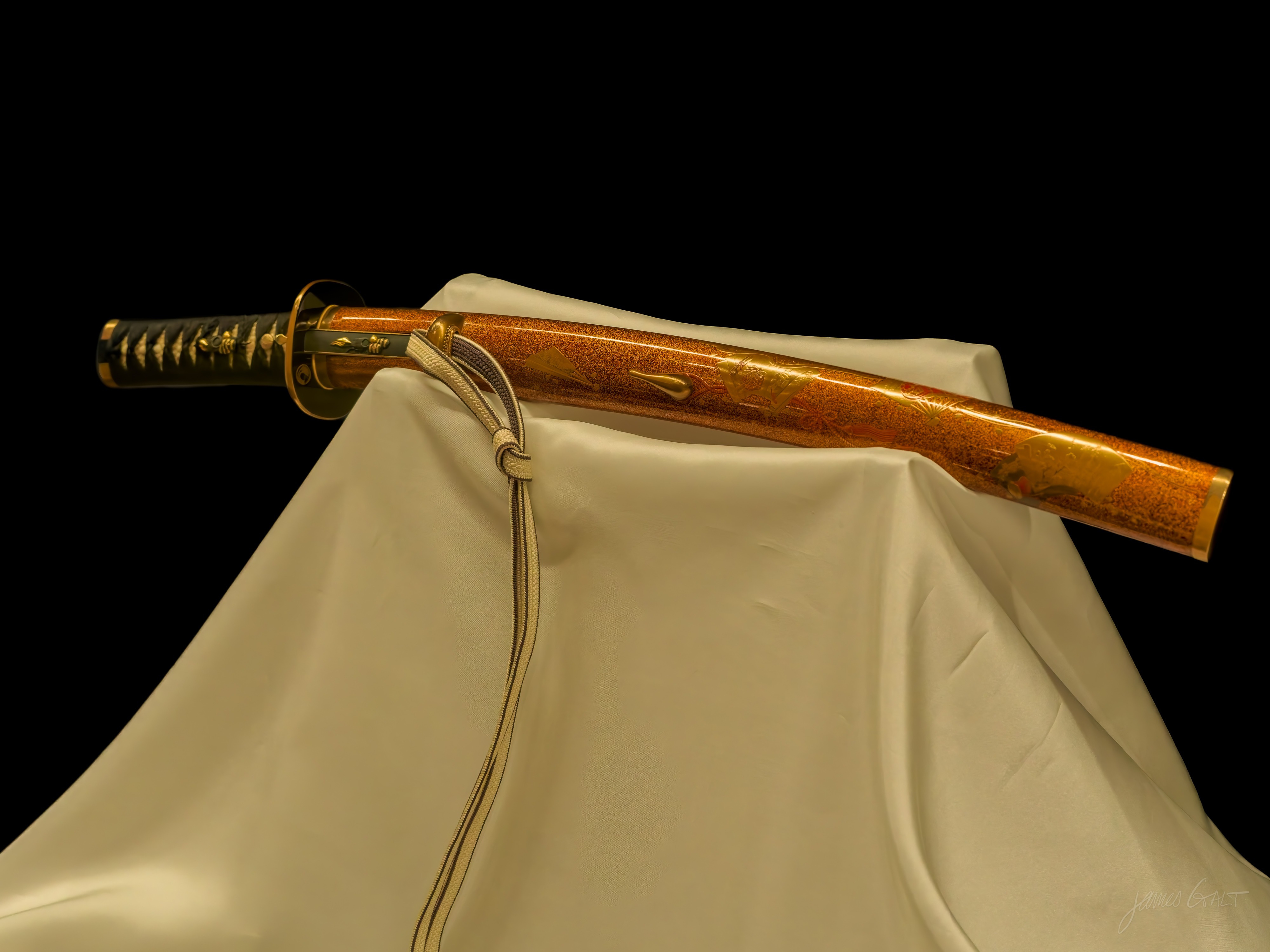A katana displayed behind glass against a black background, resting on cream silk draped over a stand. The scabbard is finished in elaborate gold lacquer, the hilt wrapped in black cord with gold fittings, and a pale cord hangs down the front of the cloth.