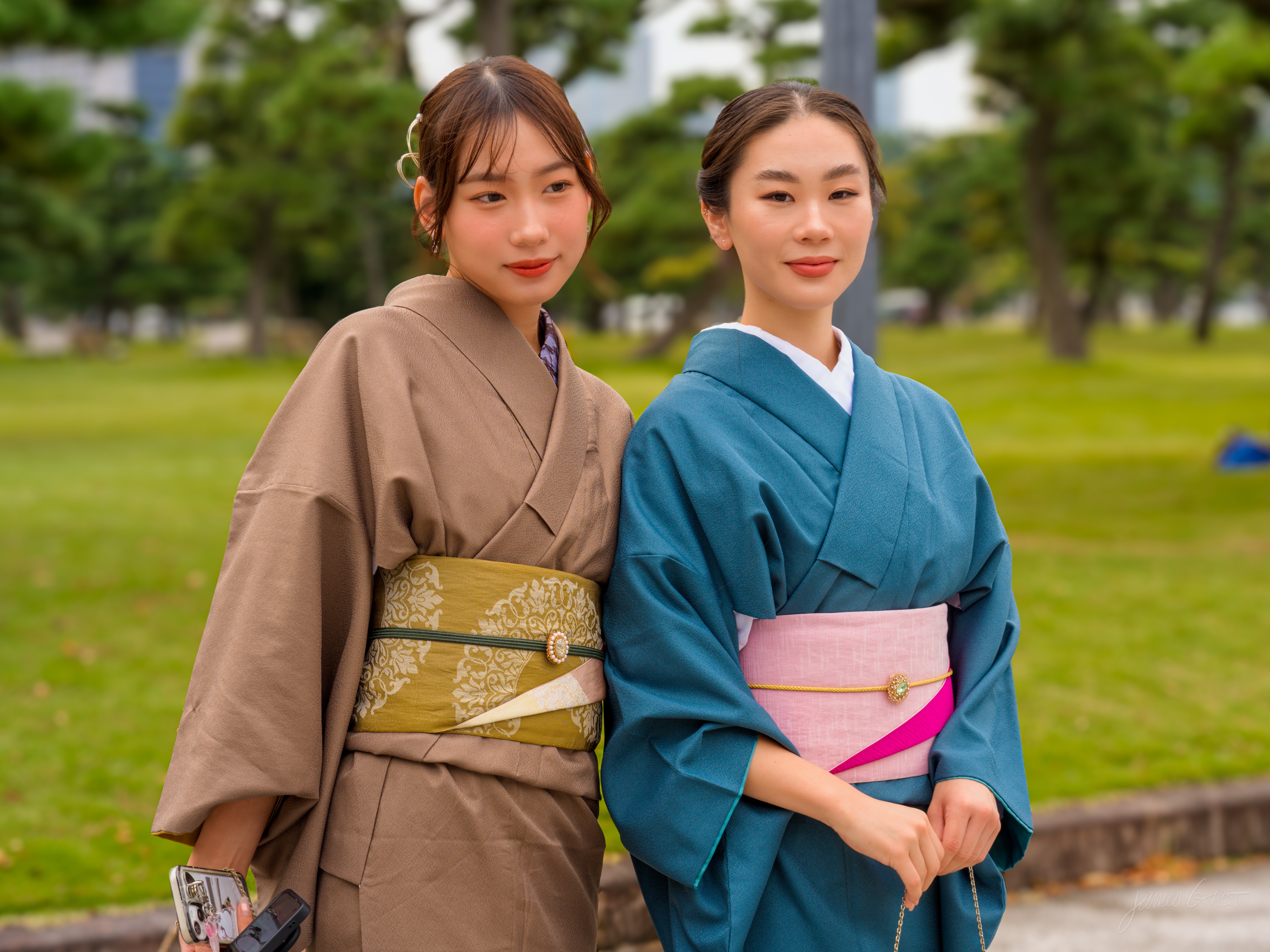 Two young women pose side by side on a lawn, both wearing kimono, one taupe with a gold sash and the other teal with a pink sash. They look toward the camera with slight smiles, green grass and pine trees softly blurred behind them. The woman on the left holds a smartphone down at her side.