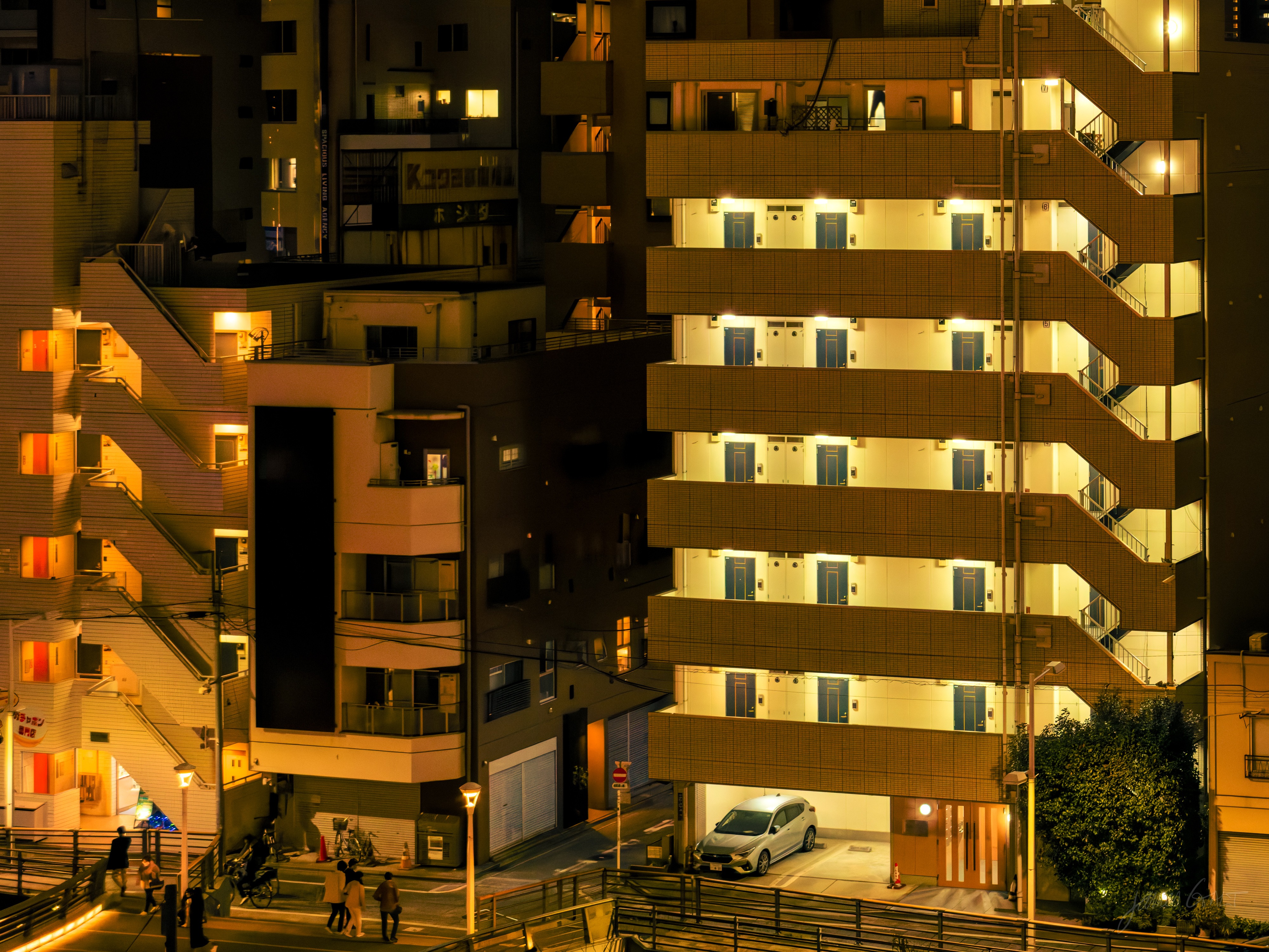 A telephoto night view of stacked apartment buildings lit warm gold, rows of external walkway balconies and a white car parked in a ground-floor bay, with a few people on a street and footbridge below.