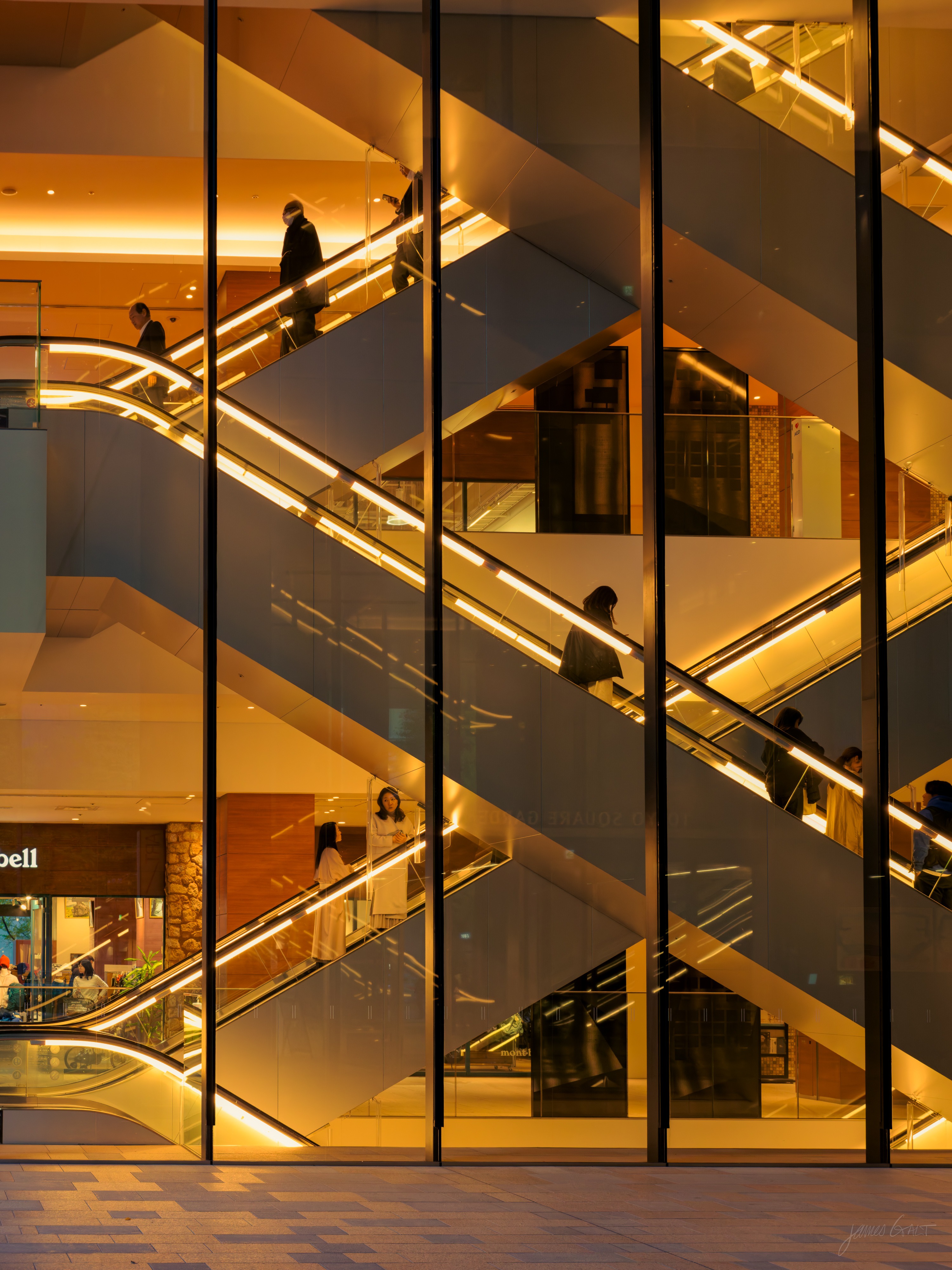 Looking through the tall glass facade of a multi-level shopping complex at criss-crossing escalators lit warm gold, several people riding them between floors, a shop unit visible at the bottom left.