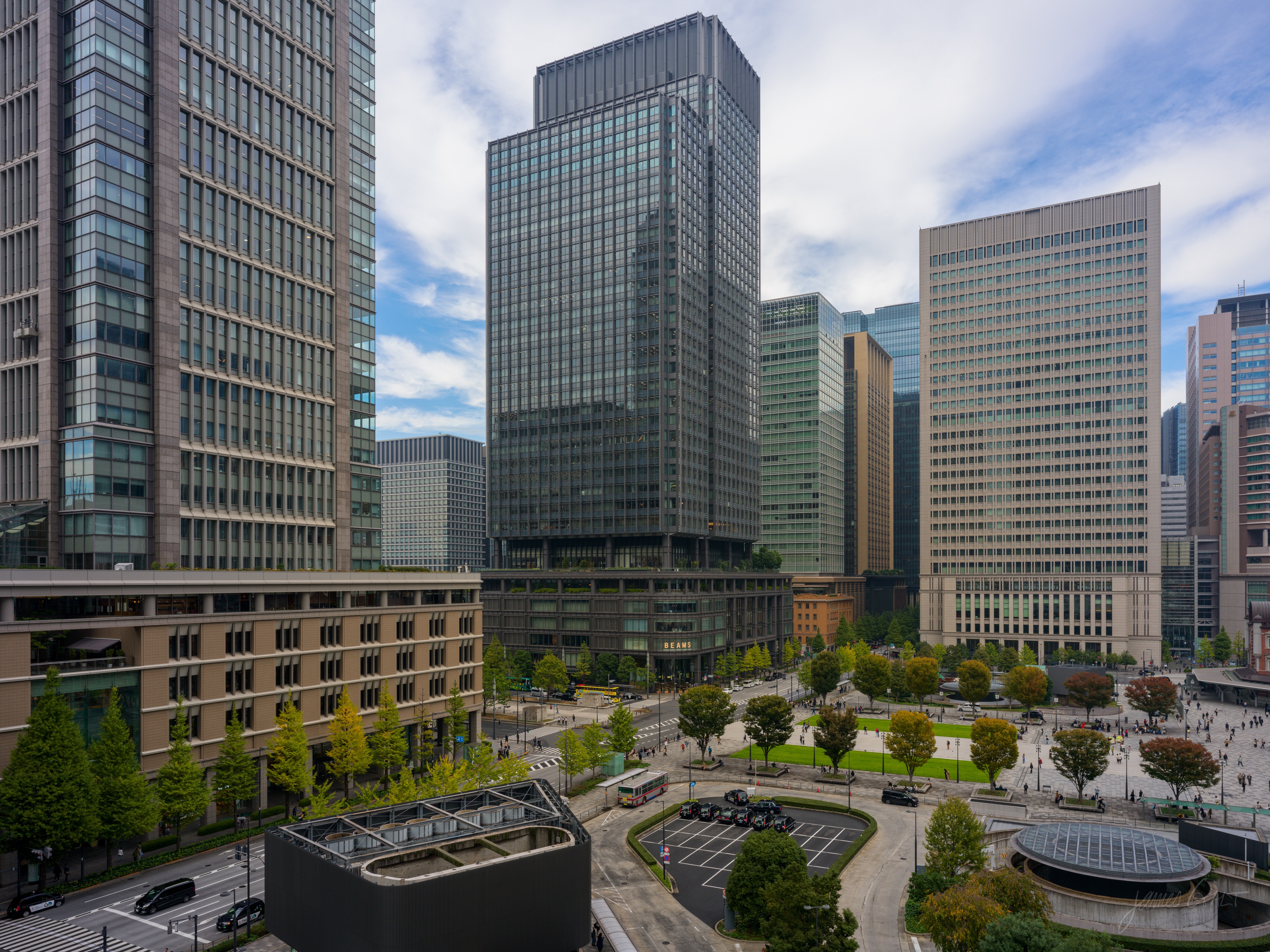 An elevated view over the Marunouchi business district: ranks of glass office towers behind a broad plaza with oval lawns and tree-lined paths, with buses and cars on the road below.