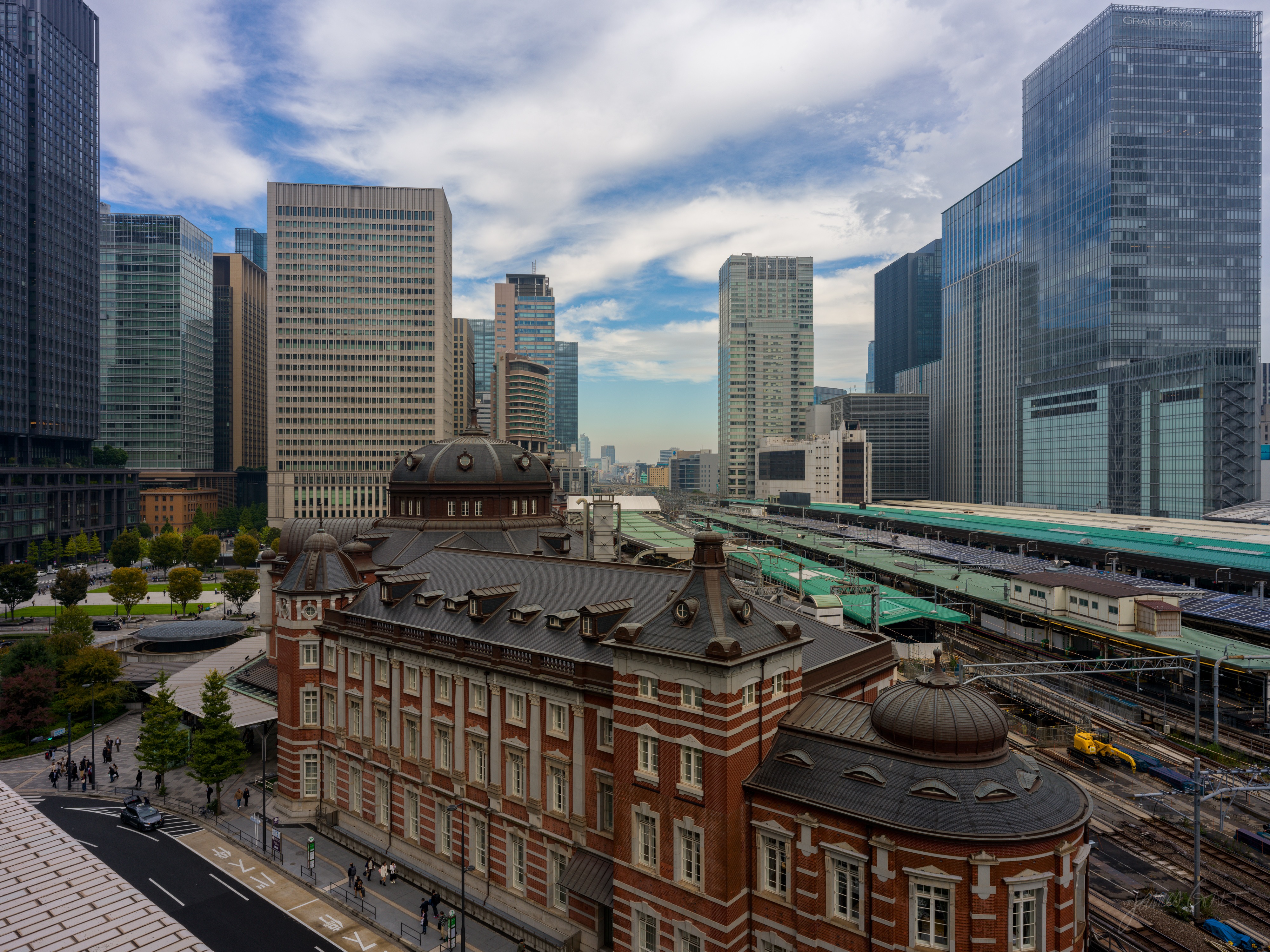 Looking down from a rooftop at the red-brick Marunouchi building of Tokyo Station with its domed roofs, the station platforms and tracks stretching away to the right and glass skyscrapers rising behind, under a blue sky with scattered cloud.