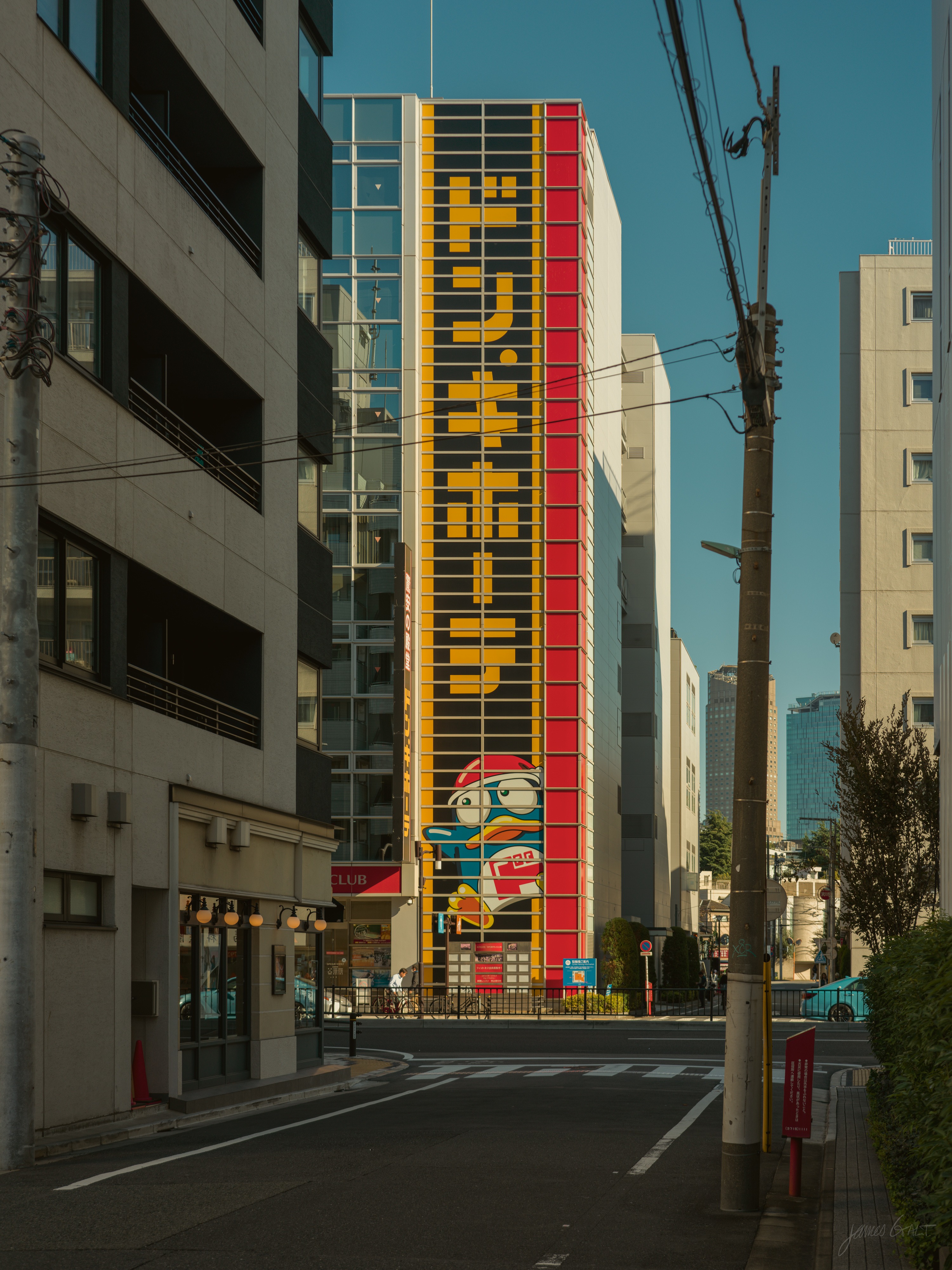 A tall, narrow Don Quijote store on a quiet morning street, its full height covered by a vertical yellow, red and black sign with a penguin mascot mural near the base. Low sun rakes across an empty intersection with power lines overhead and a red postbox at the right.