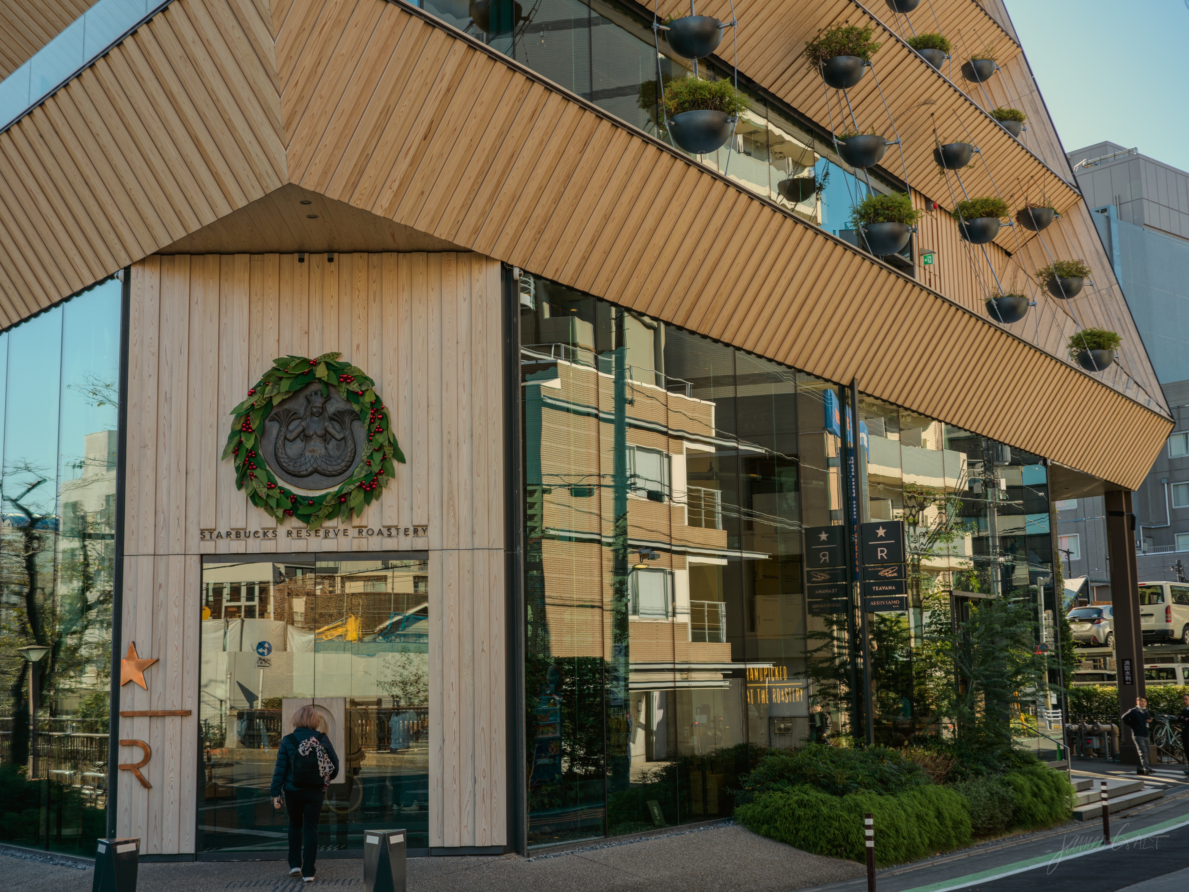 A modern building clad in angled timber battens with rows of small hanging potted plants, housing a Starbucks marked by a round green seal set in a holiday wreath. A person in dark clothing walks toward the glass entrance in soft morning light.