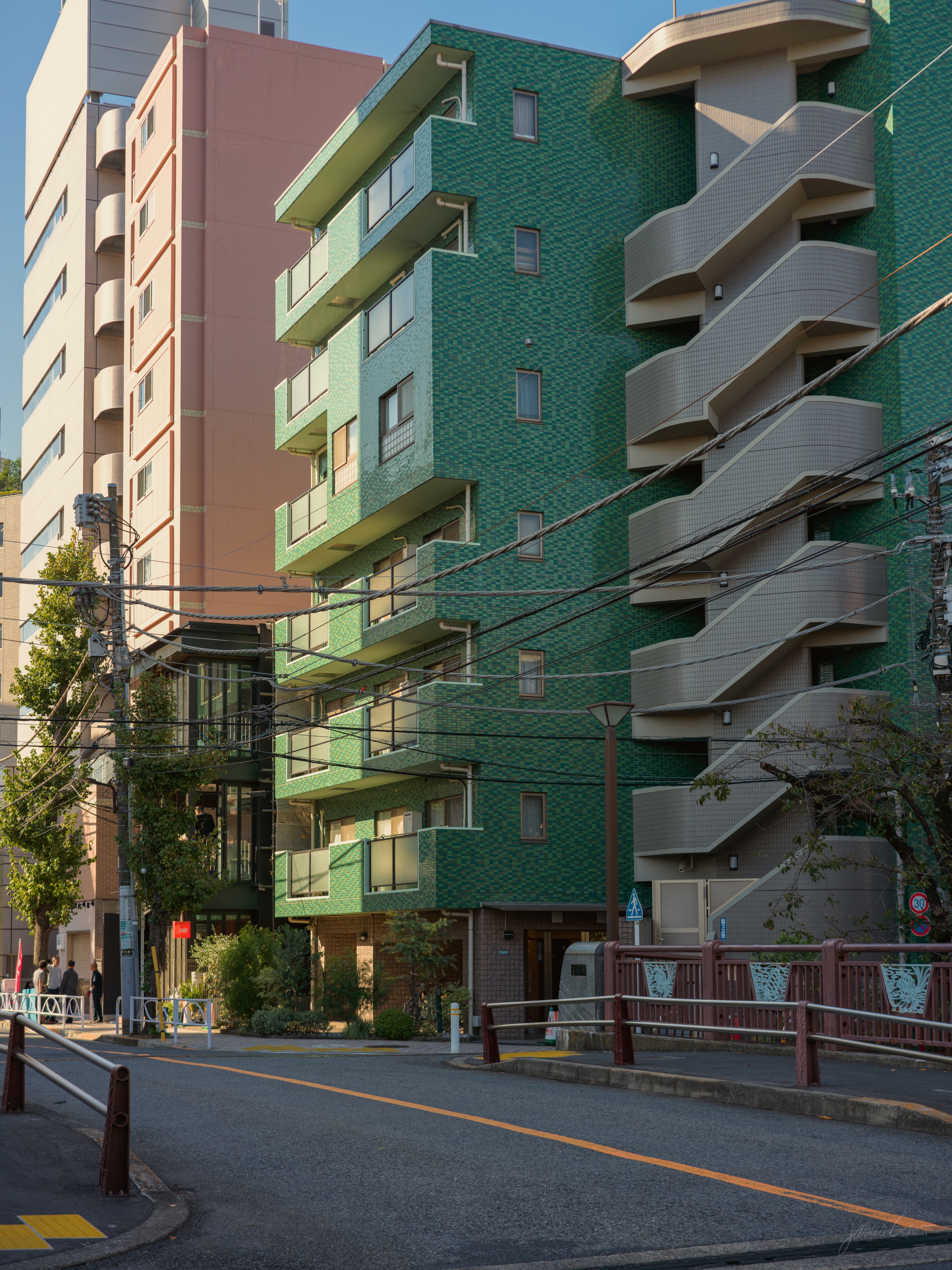 A quiet residential street in the morning, with a six-storey apartment building faced in green tile and an external brown spiral staircase tower. A small bridge with red railings crosses in the foreground and power lines run above the road.