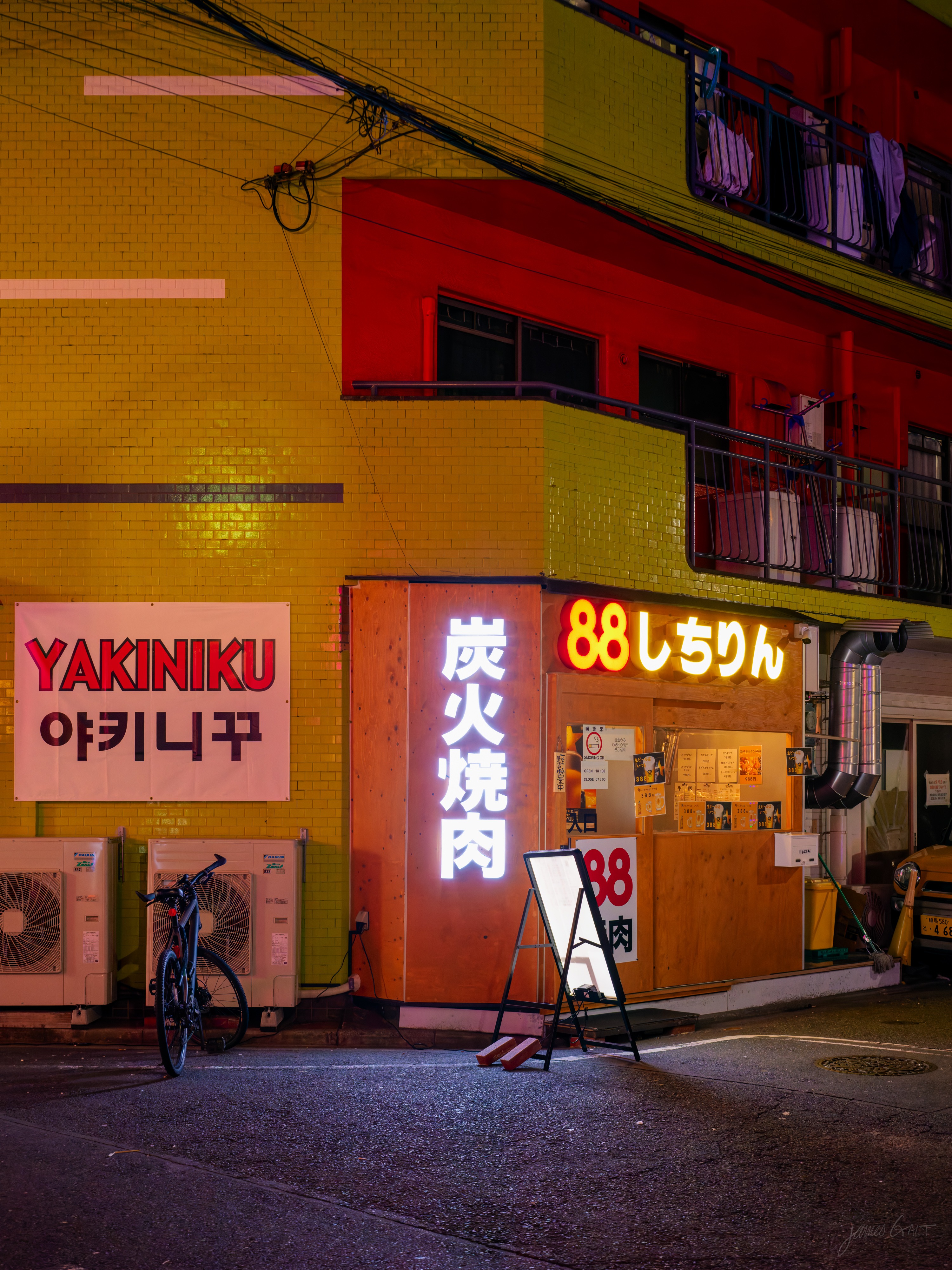 A yakiniku restaurant on a quiet street at night, its yellow-and-red tiled facade lit by neon signs reading Yakiniku and 88, with vertical red kanji for charcoal-grilled meat. A bicycle leans against air-conditioning units to the left, an A-frame menu board stands out front, and a taxi waits at the right edge.