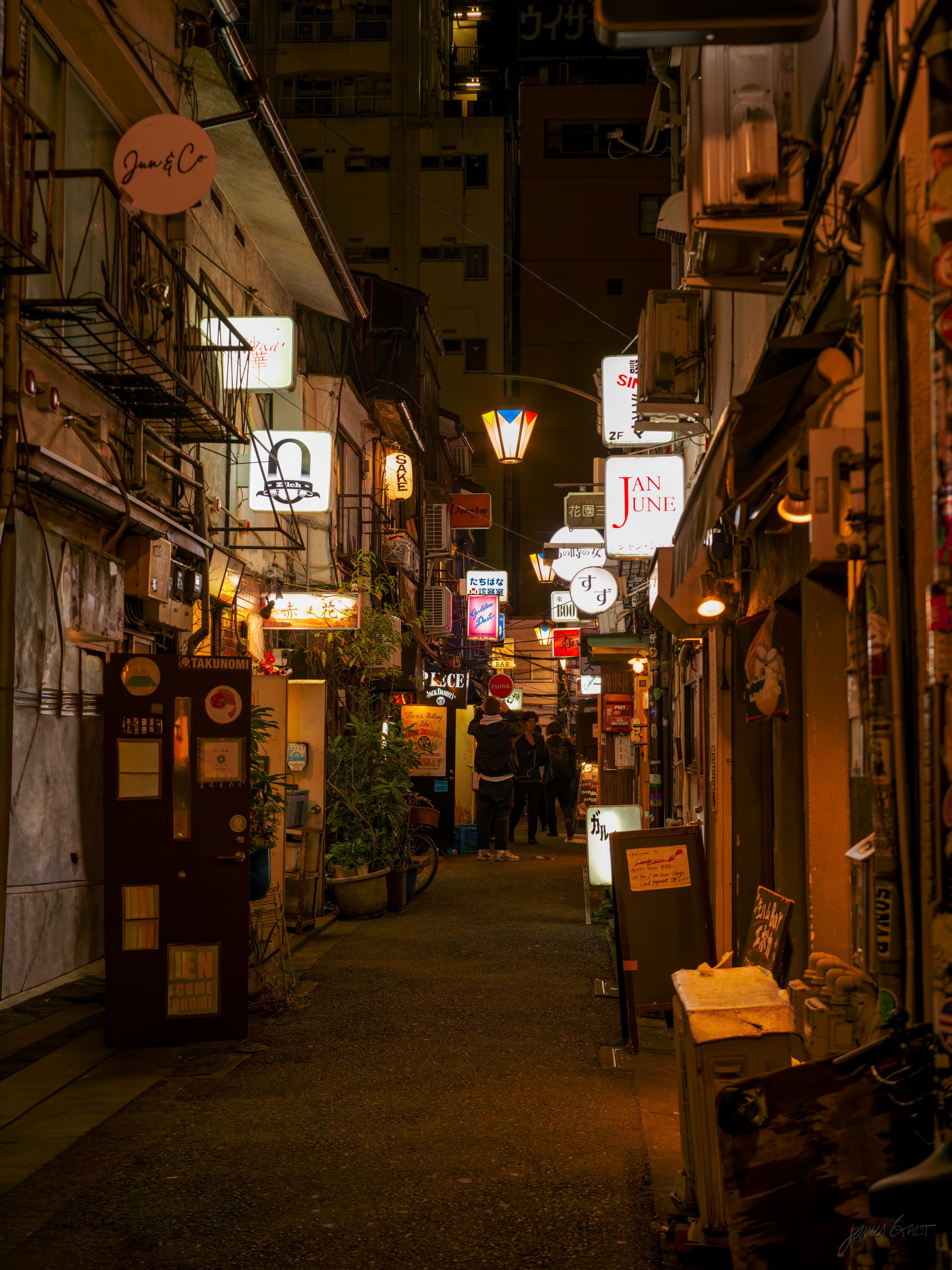 A narrow nightlife alley after dark, lined on both sides with tiny bars, stacked illuminated signs and hanging lanterns glowing warm. A small group of people stands talking in the middle distance, with a vending machine and potted plants close on the left.