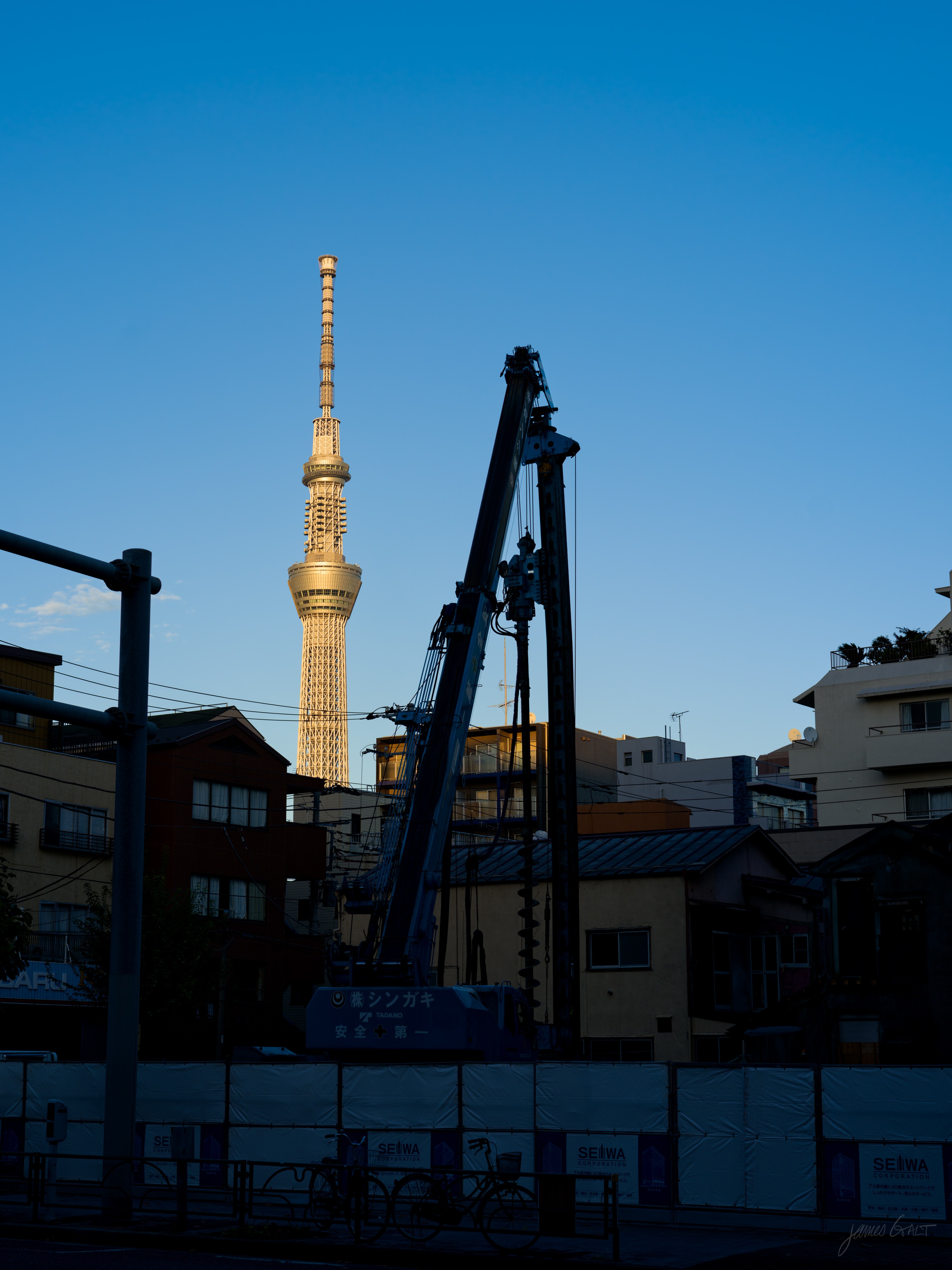 Tokyo Skytree catching low golden sunlight above a neighbourhood of low buildings, a tall black construction crane and a hoarded site in the foreground under a clear deep-blue sky.