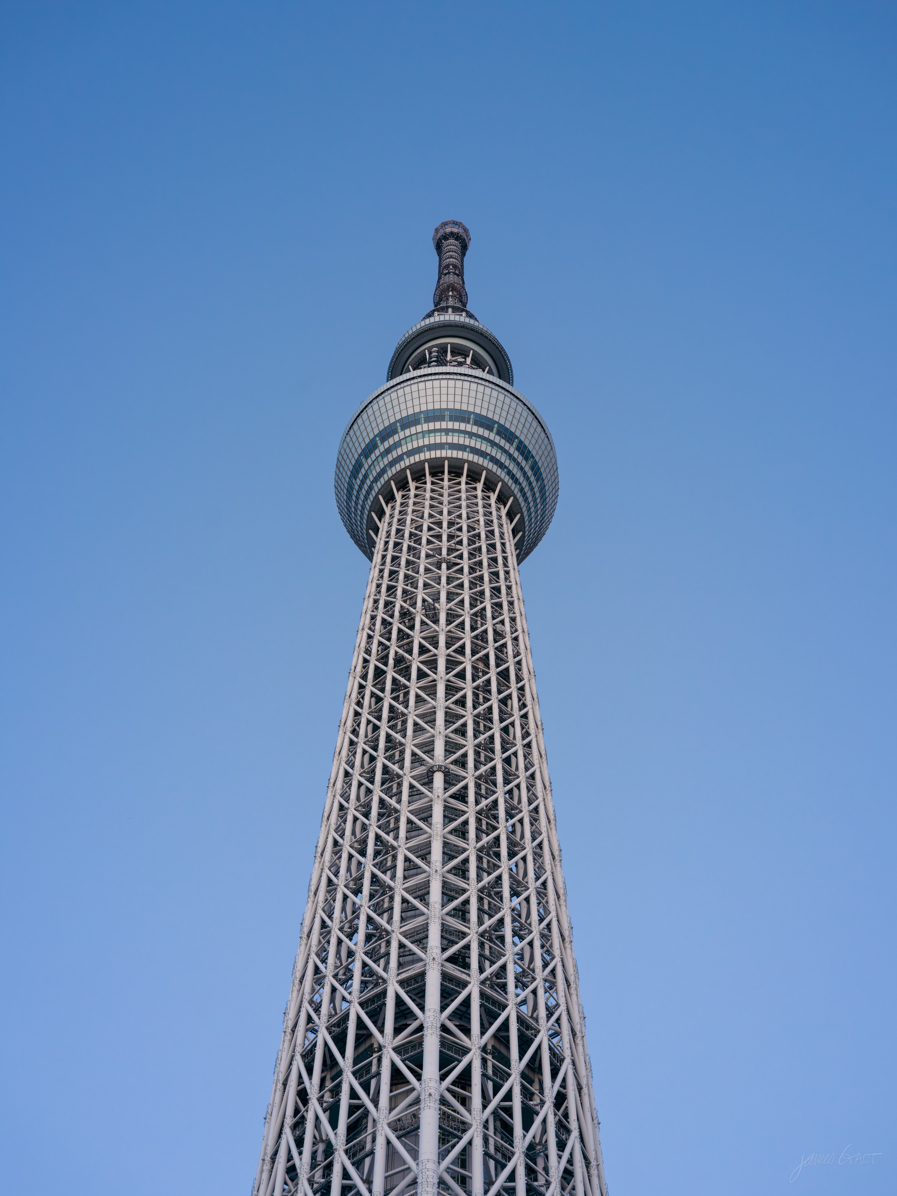Looking steeply up the Tokyo Skytree's white steel lattice to its bulged observation deck against a clear blue sky.