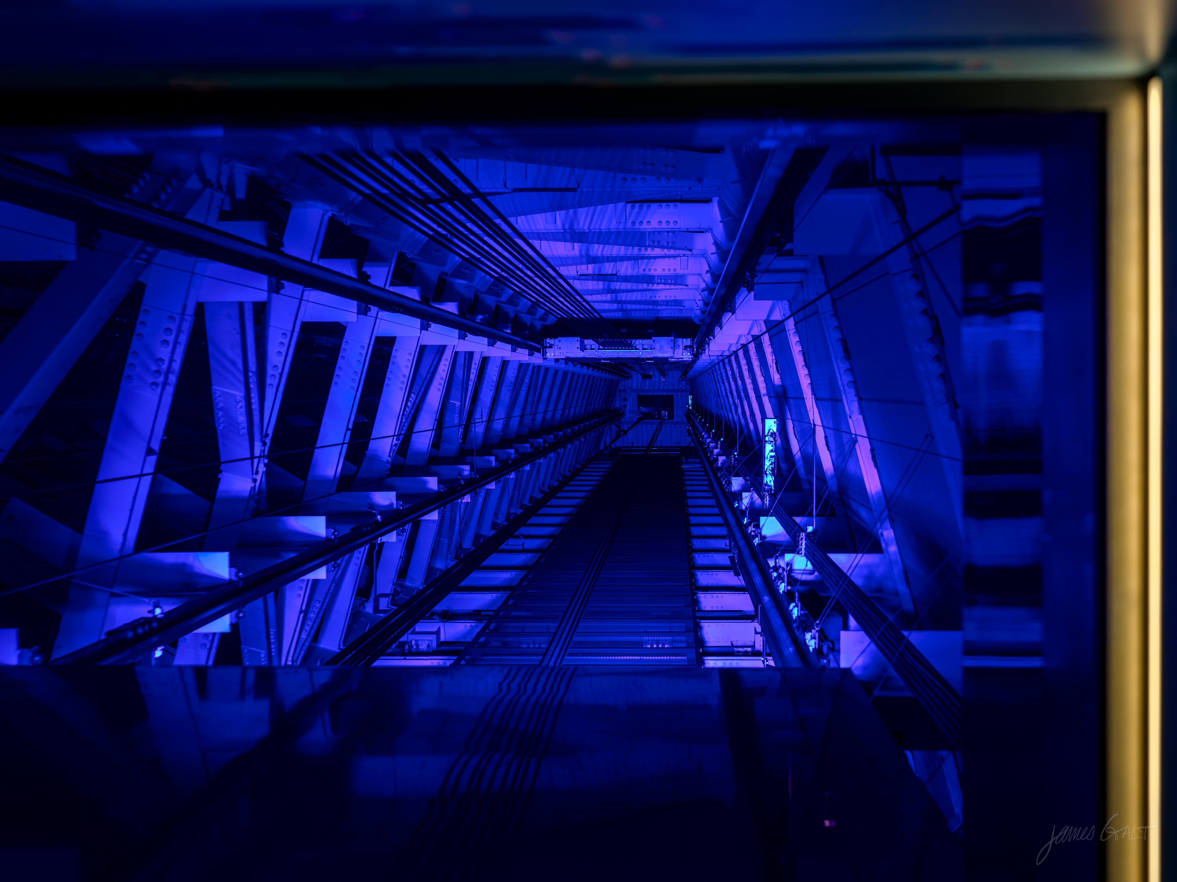 Looking up a tall internal structural shaft lit deep blue, criss-crossed steel beams receding into the distance, framed by a dark gold border.