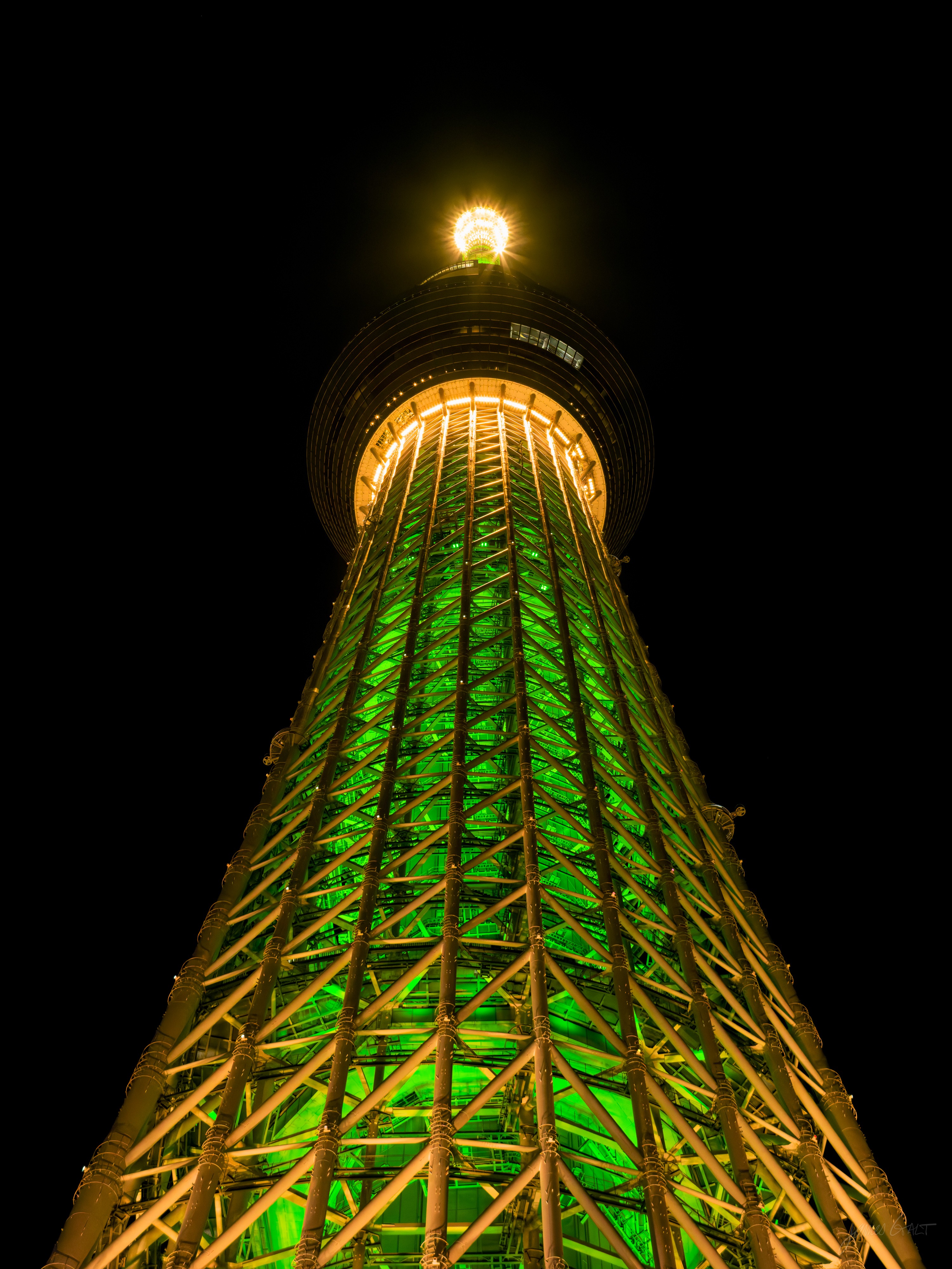 The Tokyo Skytree at night seen from its base, looking up the tapering lattice lit green and gold, a bright white light at the observation deck against a black sky.