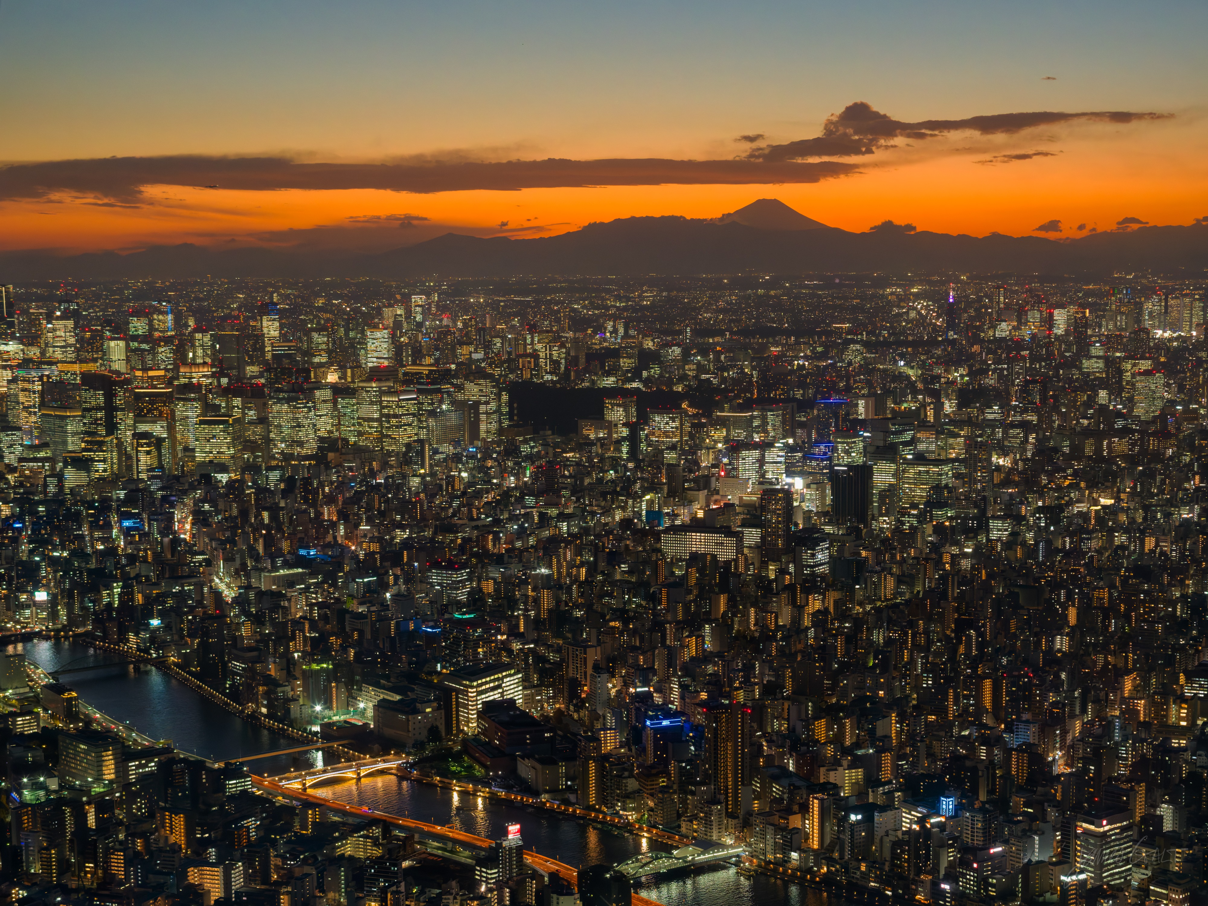 A high wide view over Tokyo at sunset, the silhouette of Mount Fuji on a glowing orange horizon, the dense city below with lights coming on and the Sumida River winding through it crossed by lit bridges.