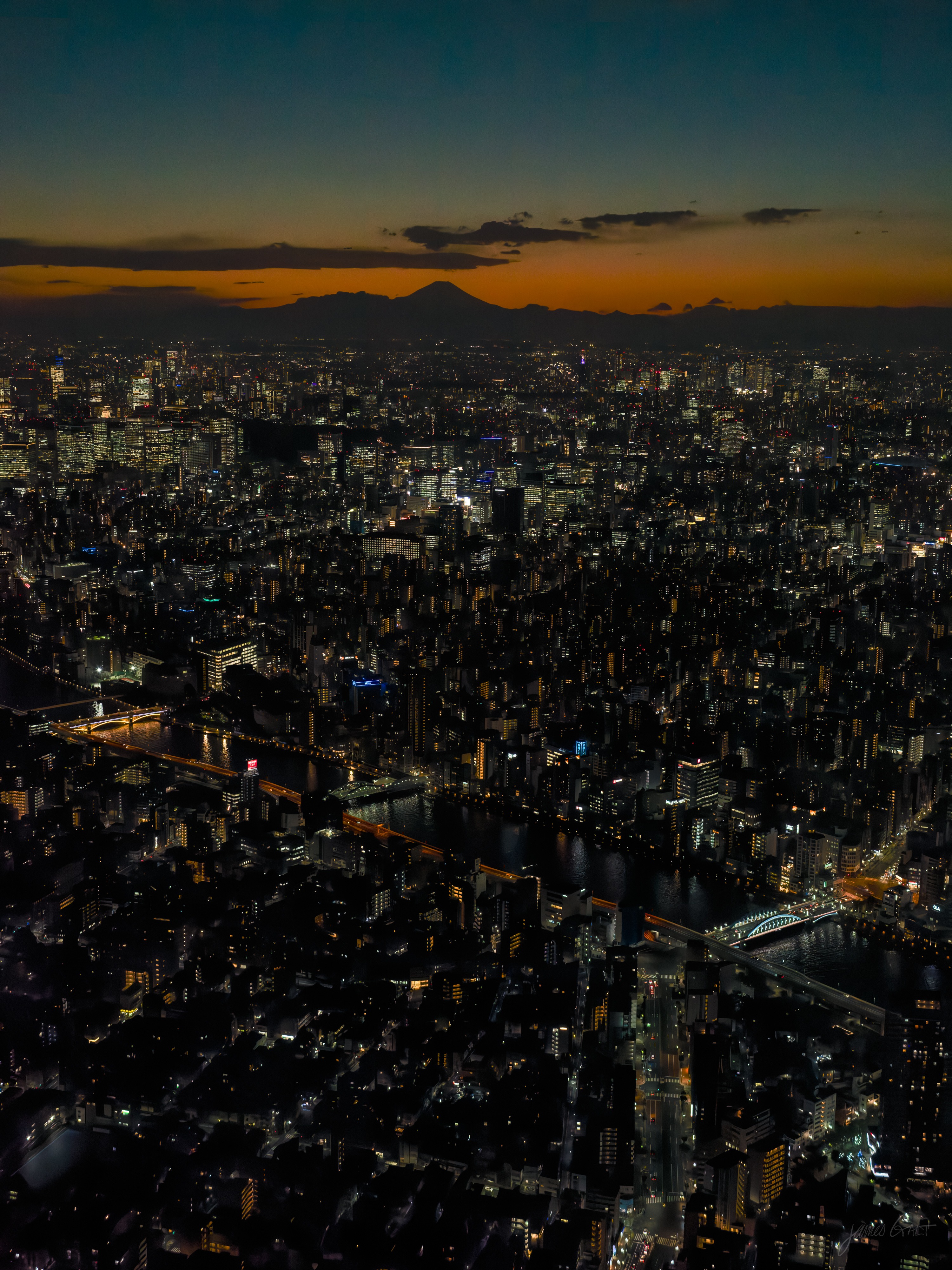 A vertical high view over Tokyo at dusk, Mount Fuji low on the orange horizon, the darkened city stretching below dotted with lights, the Sumida River and its bridges leading toward the skyline.