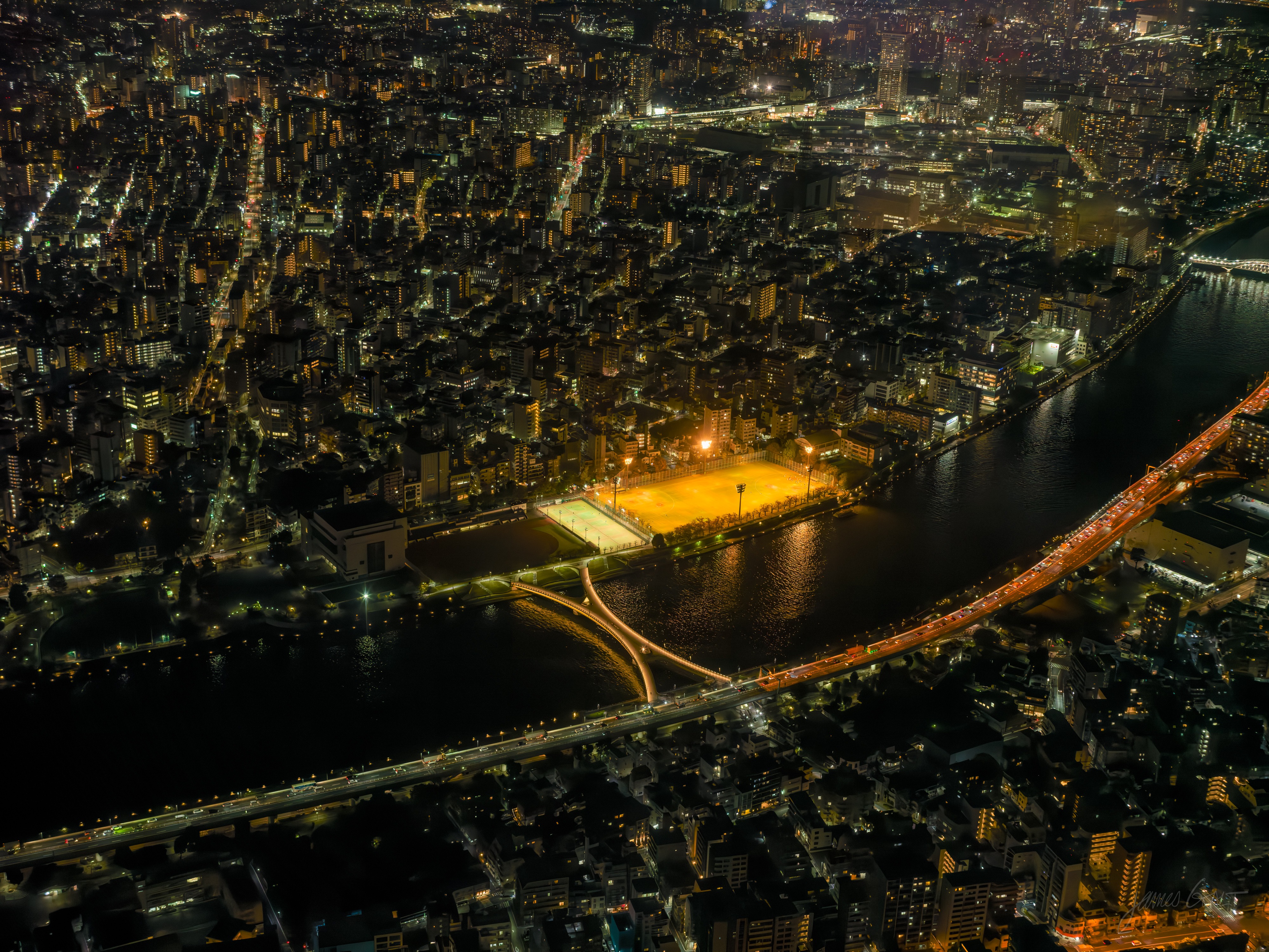 A night view from high above Tokyo: a single brightly floodlit sports field glowing orange beside the dark Sumida River, an expressway bridge picked out in light crossing the water, the city spread out around it.