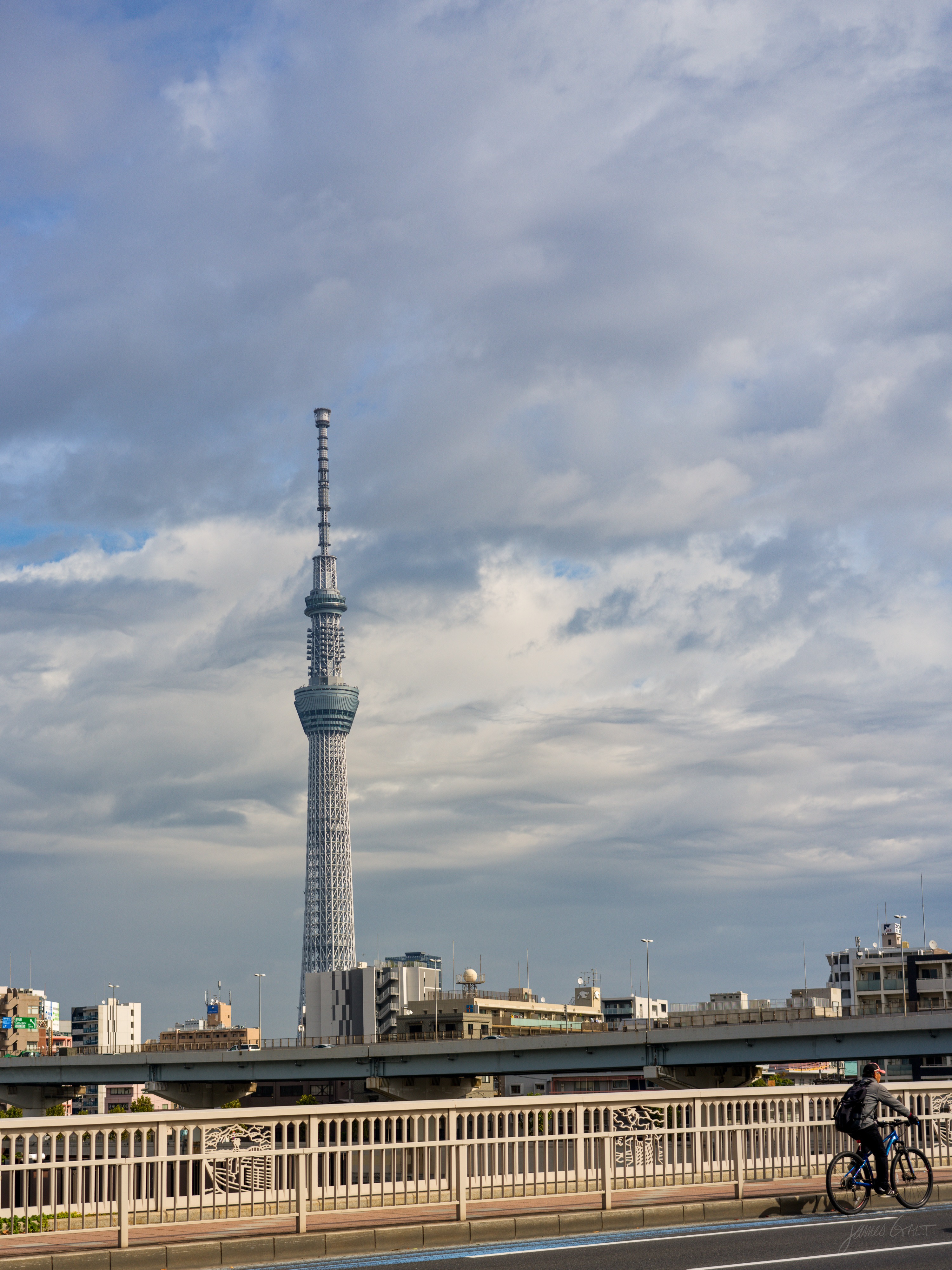Tokyo Skytree rising far above a band of low-rise buildings under a wide sky of broken cloud. A pale bridge railing runs across the foreground, and a cyclist with a backpack rides along the right-hand edge.