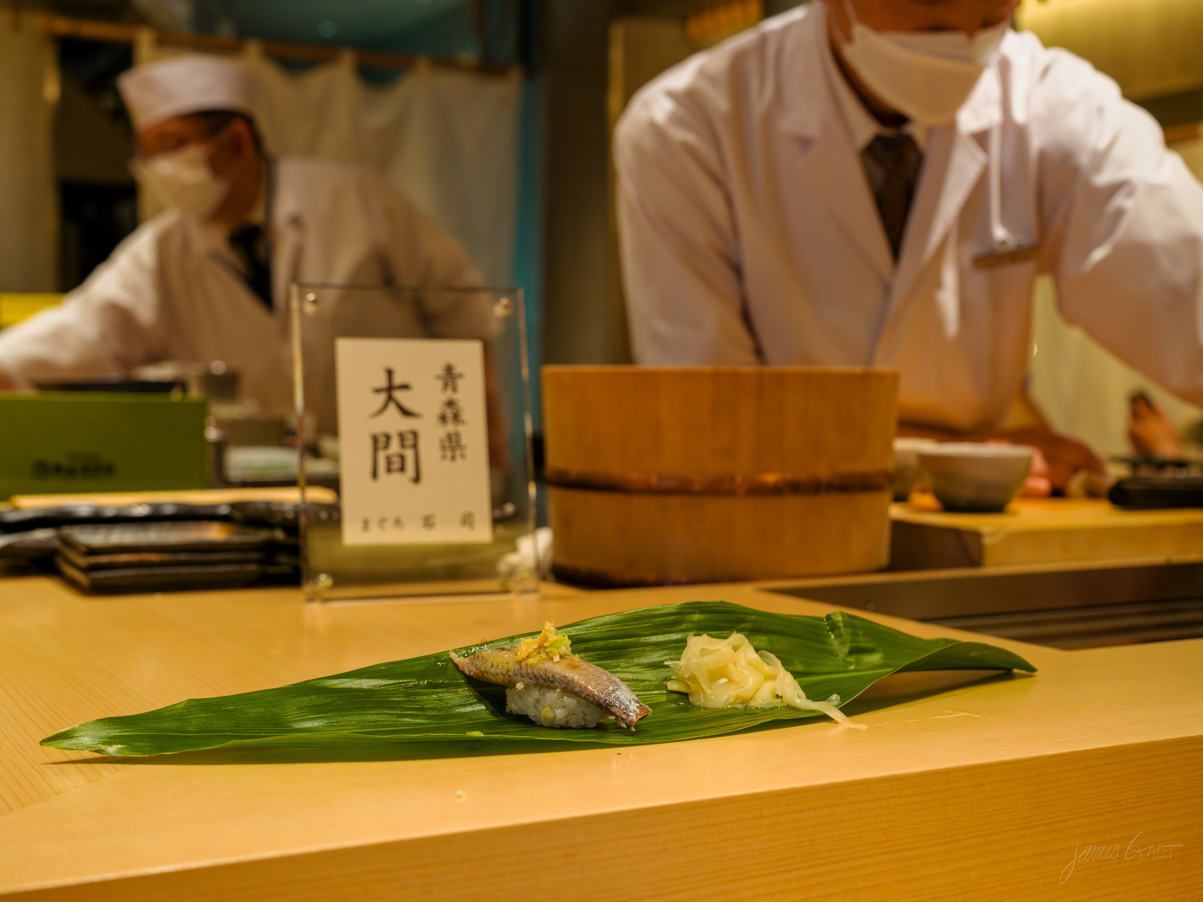 Across a pale wood sushi counter, a single piece of nigiri topped with a silver-skinned fish, grated ginger and chopped scallion, sits on a green bamboo leaf beside a small mound of pickled ginger. Behind it, two masked chefs in white coats work near a wooden rice tub and an acrylic stand holding a sign that reads Aomori, Ōma.