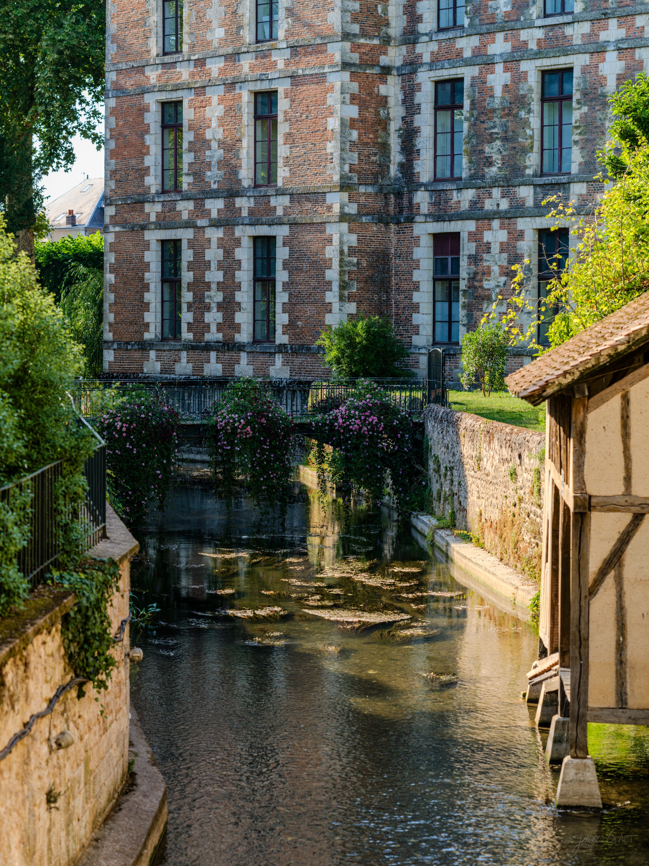A half-timbered and stone-checkered house reflected in a still canal, climbing roses on the wall, a timber-framed structure to the right.