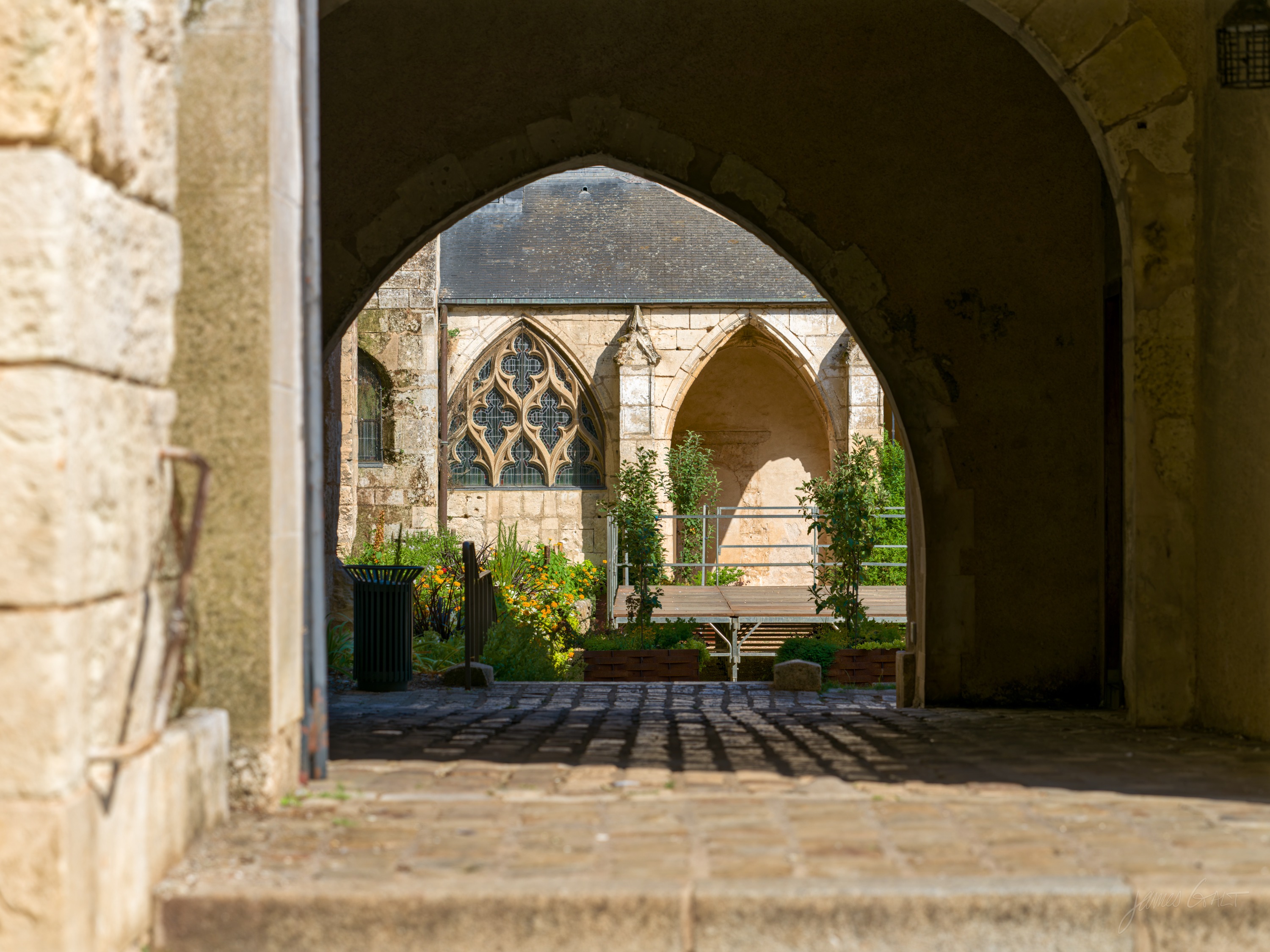 View through a pointed stone archway into a cloister courtyard, with Gothic tracery windows and raised planting beds beyond.