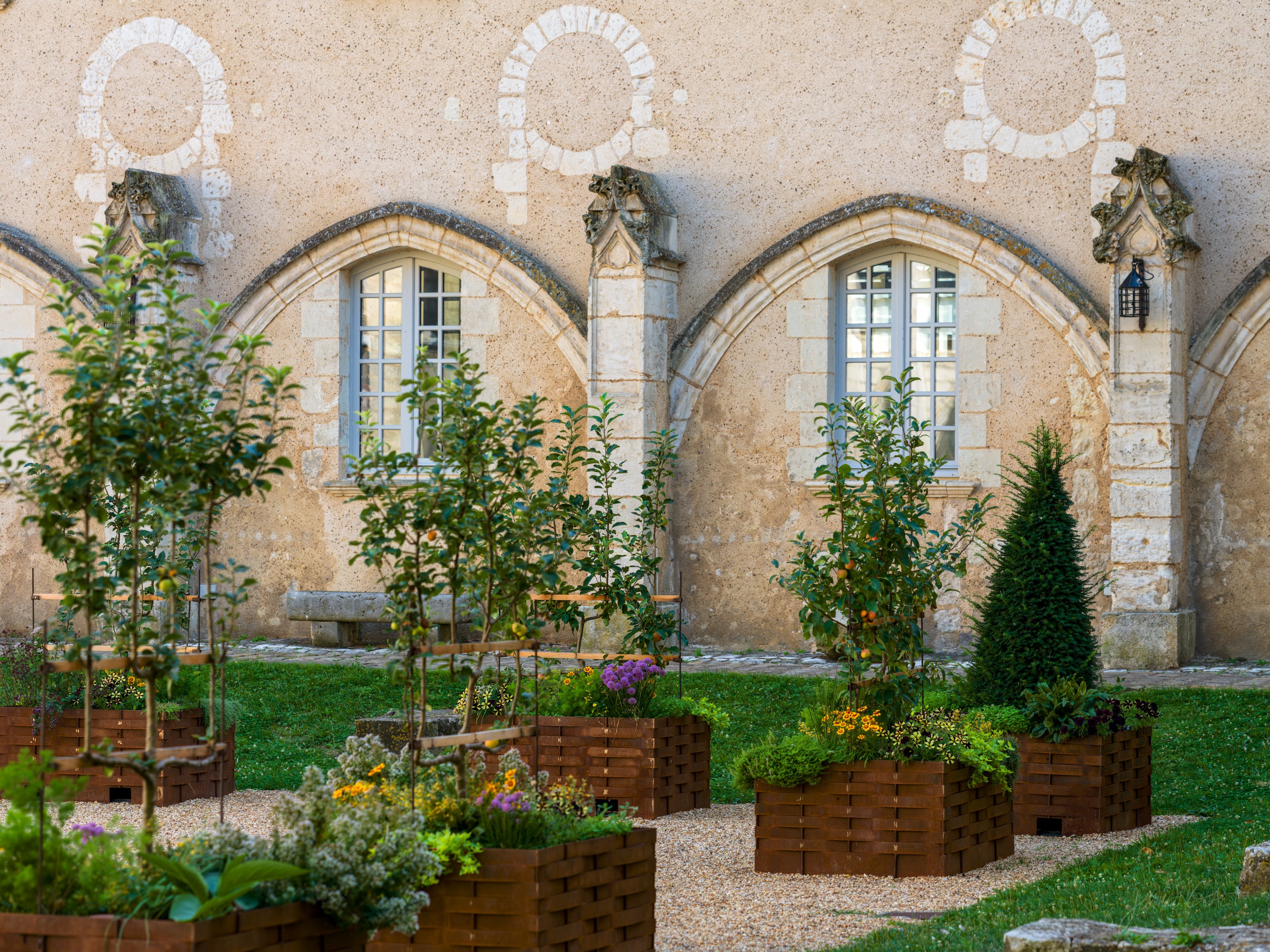 A cloister garden of woven-willow raised beds on gravel, set against a stone wall of blind Gothic arcading and mullioned windows.