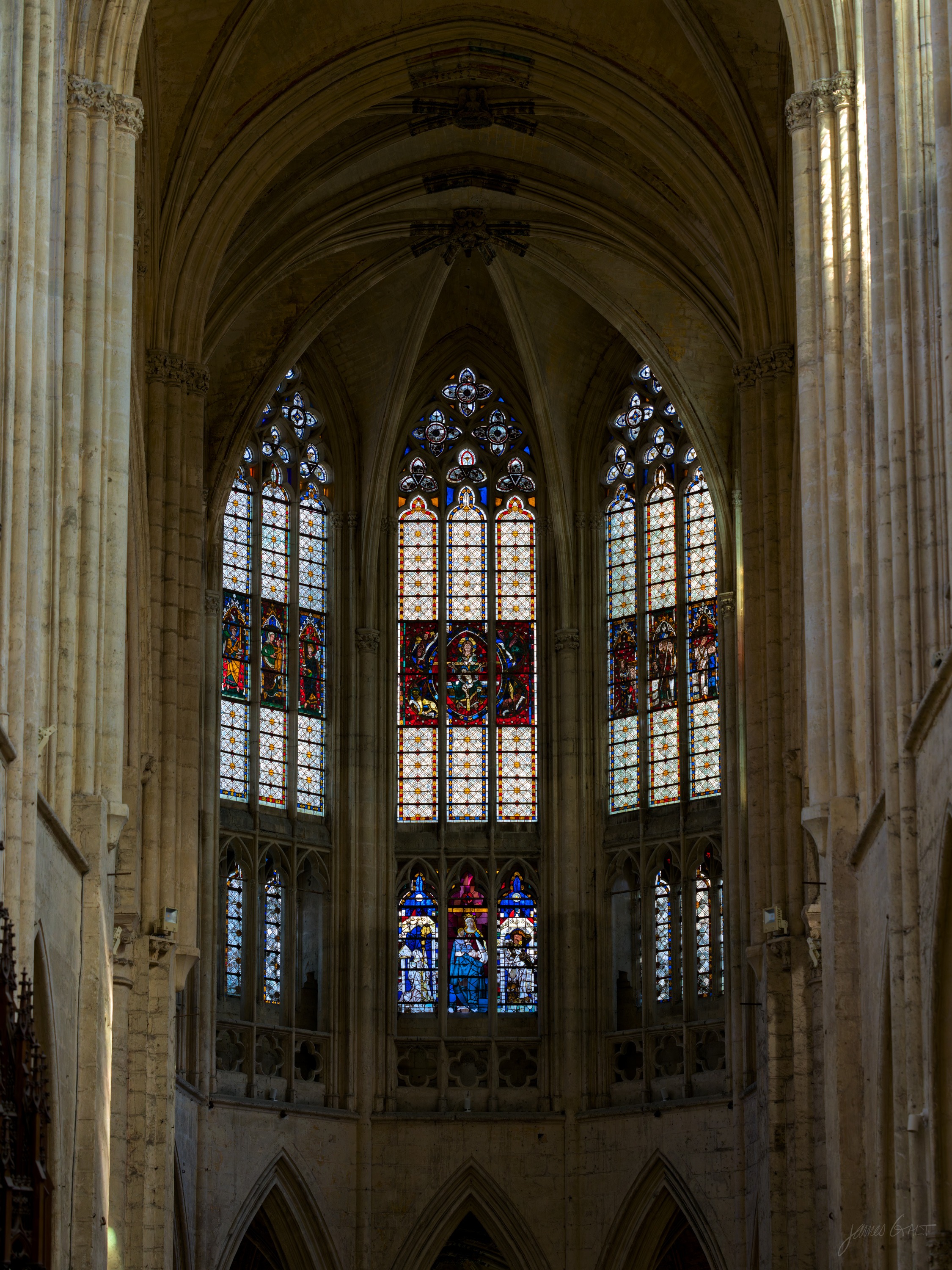 The interior of a Gothic church looking toward the apse, tall clustered columns rising to a vaulted ceiling, stained-glass windows lit at the far end.