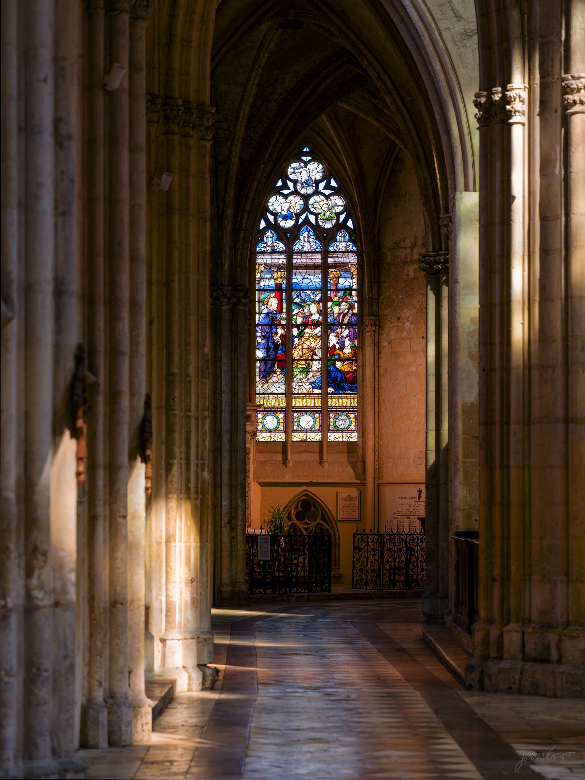 A side aisle of a Gothic church, a tall stained-glass window glowing at the end and a pool of coloured light on the stone floor.