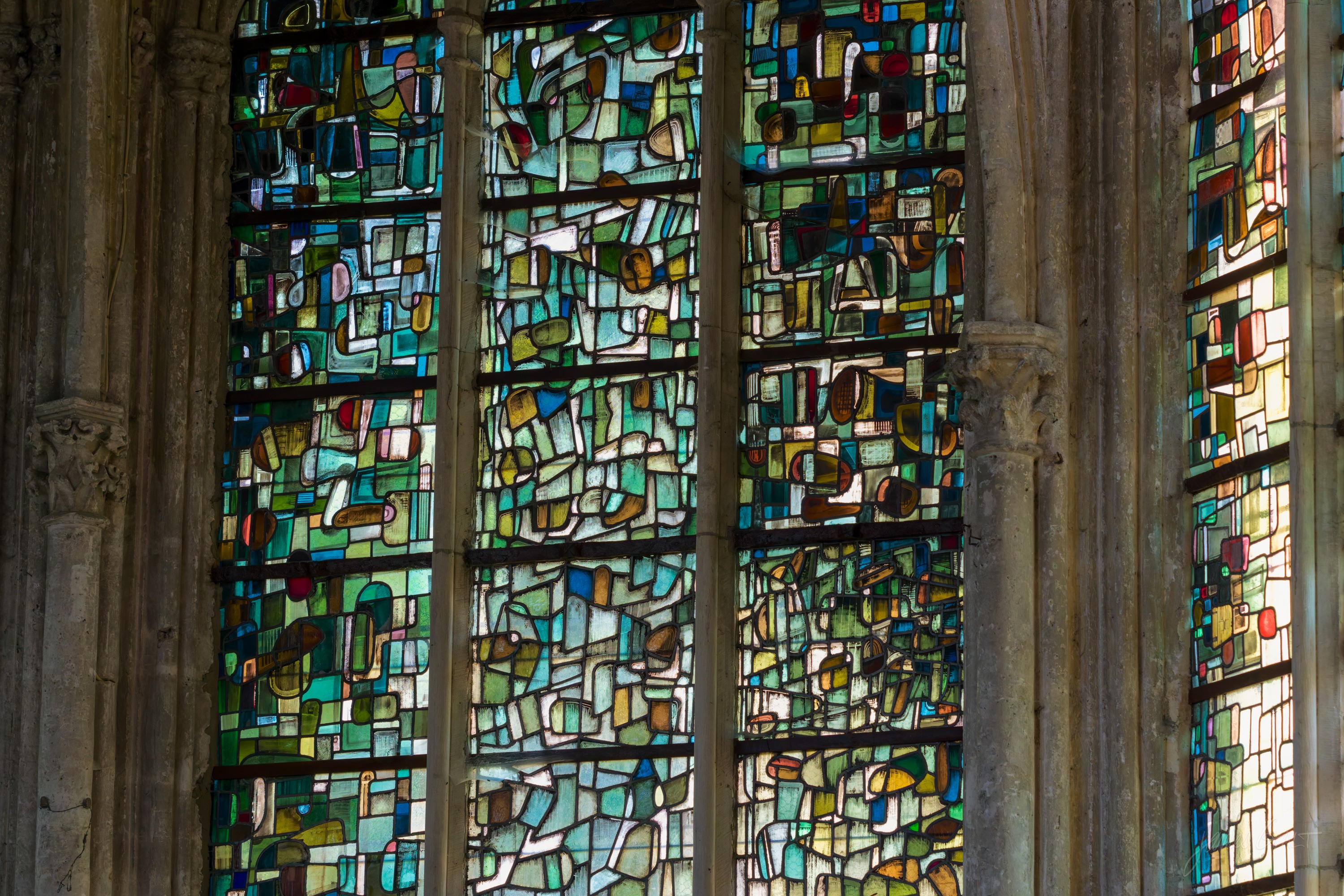 A modern abstract stained-glass window in greens, blues and browns, set within Gothic stone mullions.