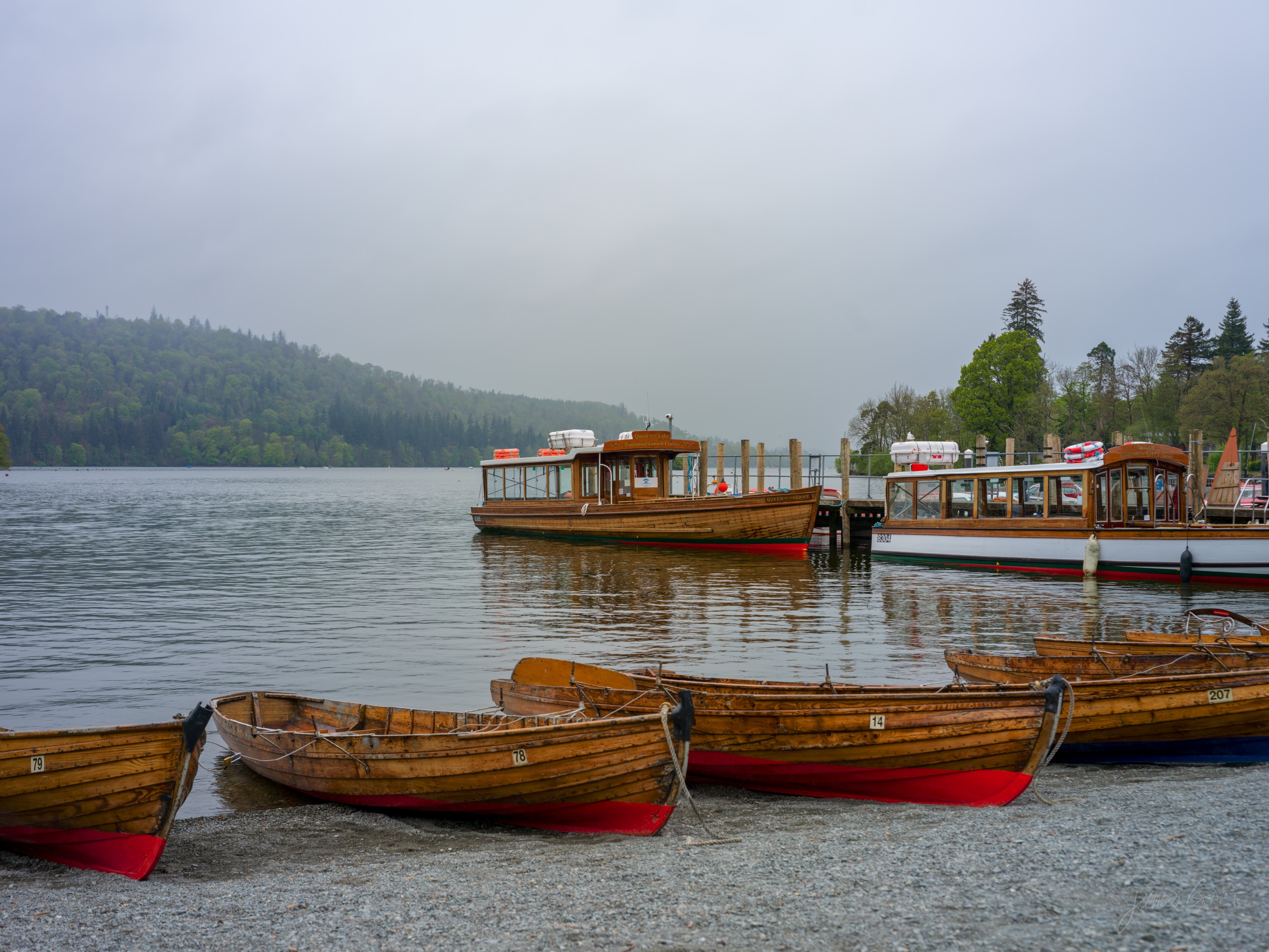 Several wooden rowing boats with red-painted keels drawn up on a shingle shore, two larger wooden lake launches moored at a jetty, wooded hills fading into rain behind.