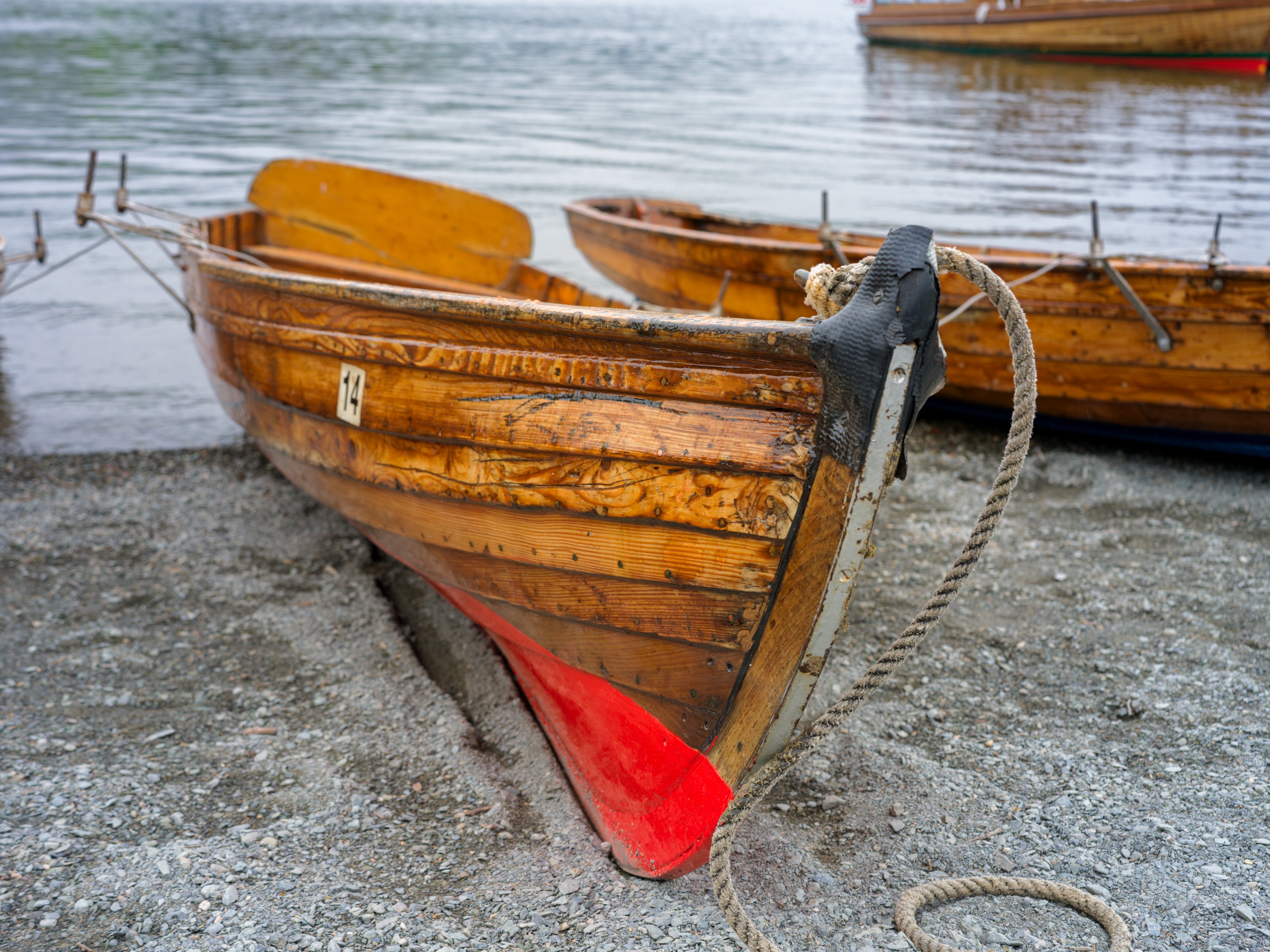 Close view of the bow of a varnished wooden rowing boat on grey shingle, a mooring rope looped from its stem, a red-painted keel beneath, other boats behind.
