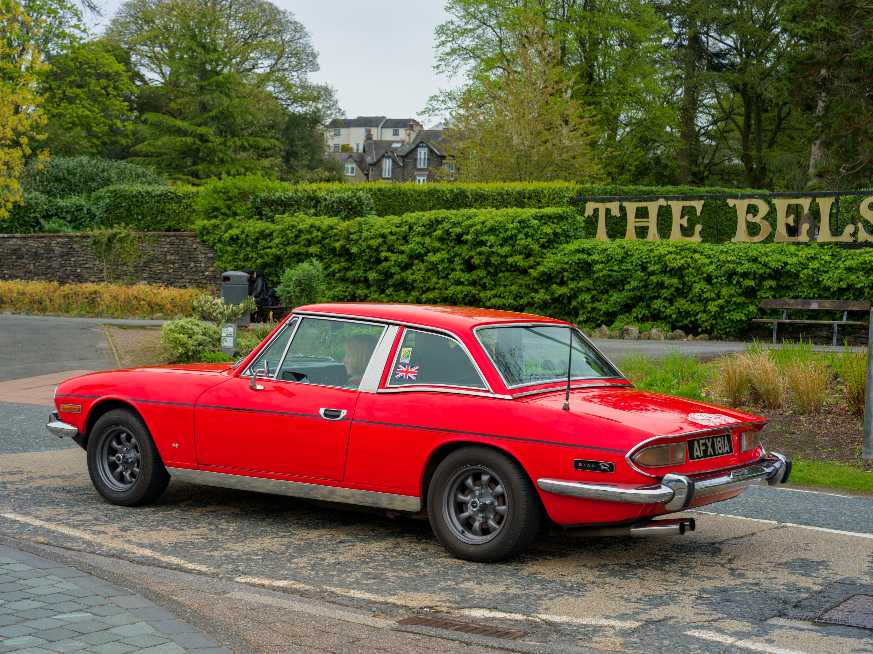 A red Triumph Stag parked on a stone-paved street beside a clipped green hedge, gold lettering reading THE BELS on the wall behind.
