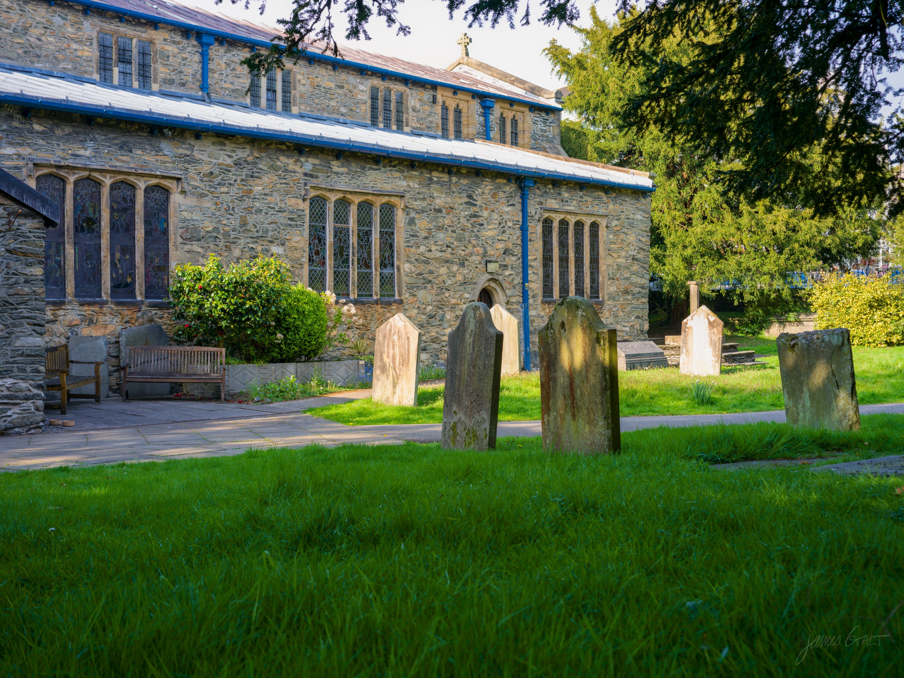 A stone-built church with lancet windows framed in blue paint, lit warm by low evening sun, three weathered headstones leaning on bright green grass in the foreground, a dark yew to the right.
