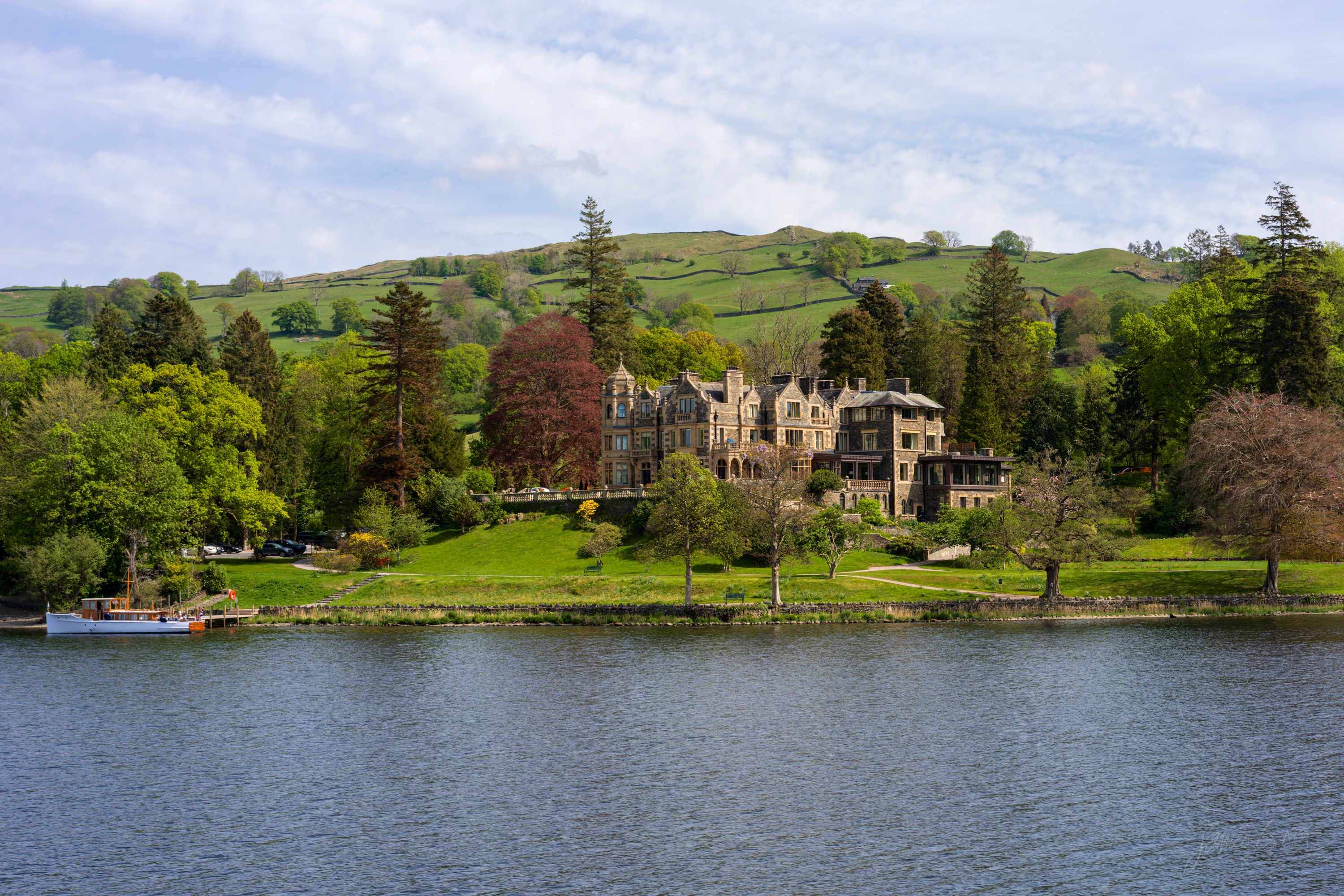 A large stone Victorian house with gables set in lawns and trees on the far shore of a lake, green fields rising to a low ridge behind, calm water in front.