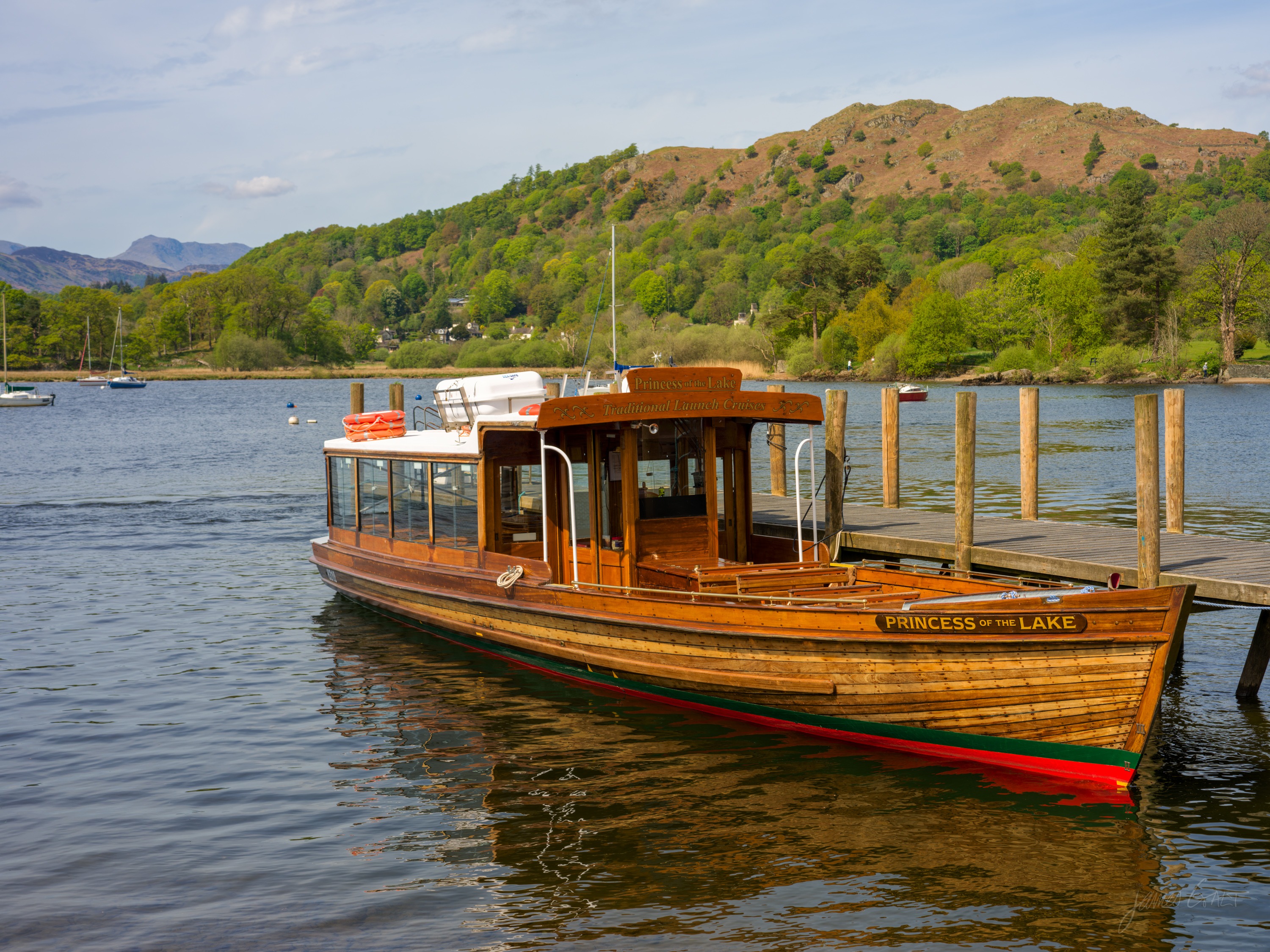 A varnished wooden passenger launch named Princess of the Lake moored at a jetty on calm water, a bracken fell and wooded shore behind under blue sky.