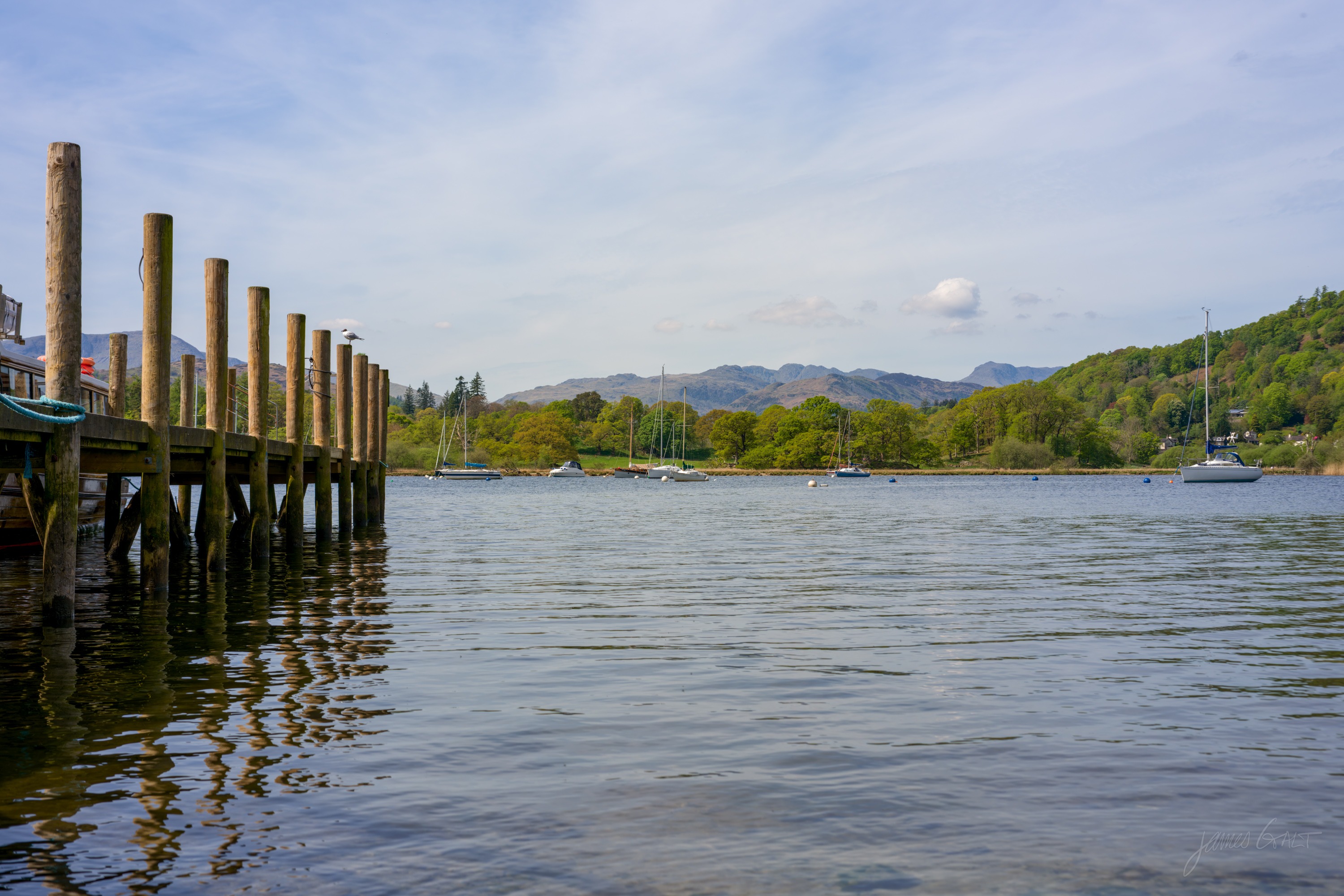A wooden jetty on tall pilings seen low from the waterline, moored yachts on the lake beyond and hazy fells under a bright sky.