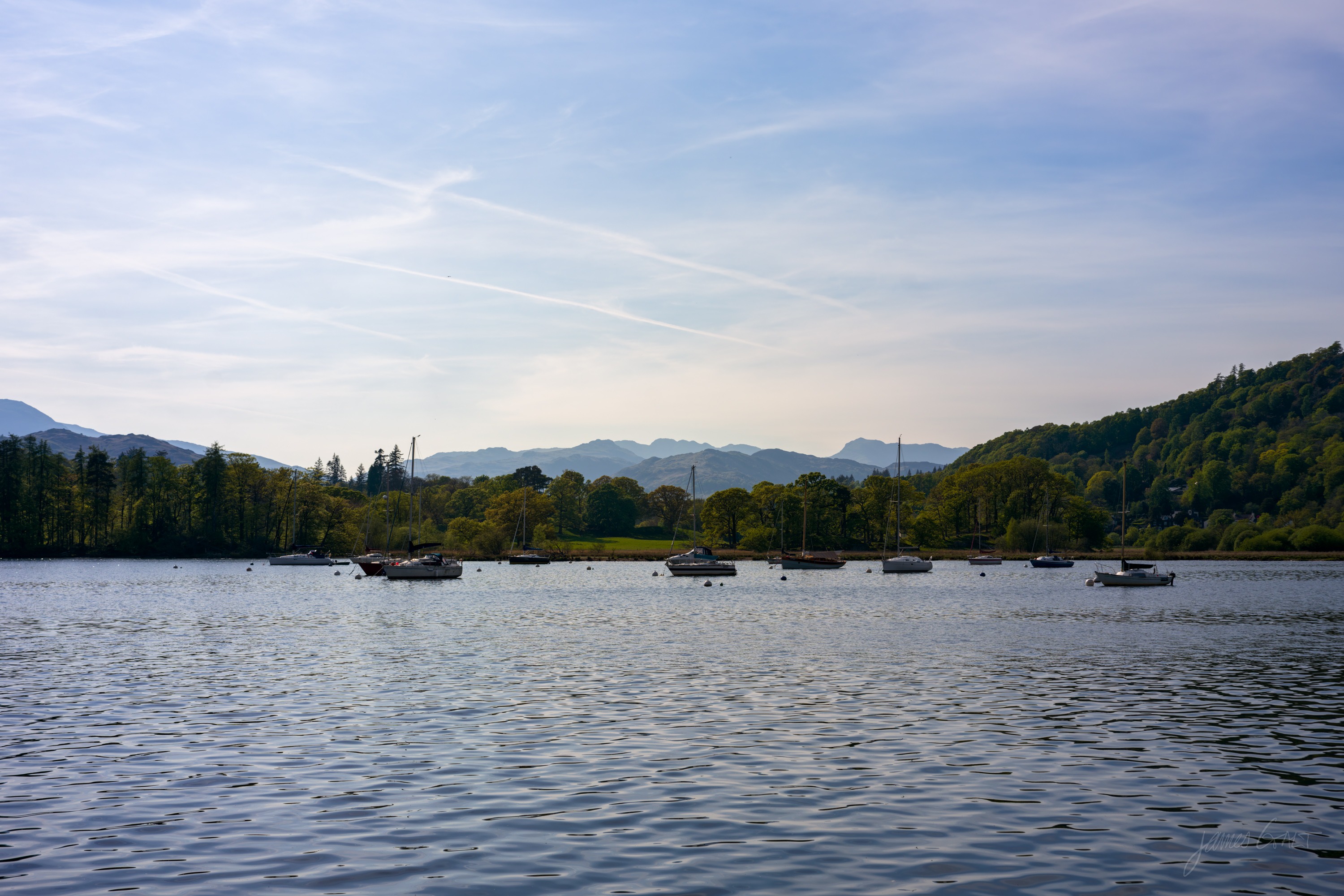 A wide calm stretch of Windermere with small moored boats, wooded shores on both sides, distant fells and aircraft contrails crossing a pale blue sky.