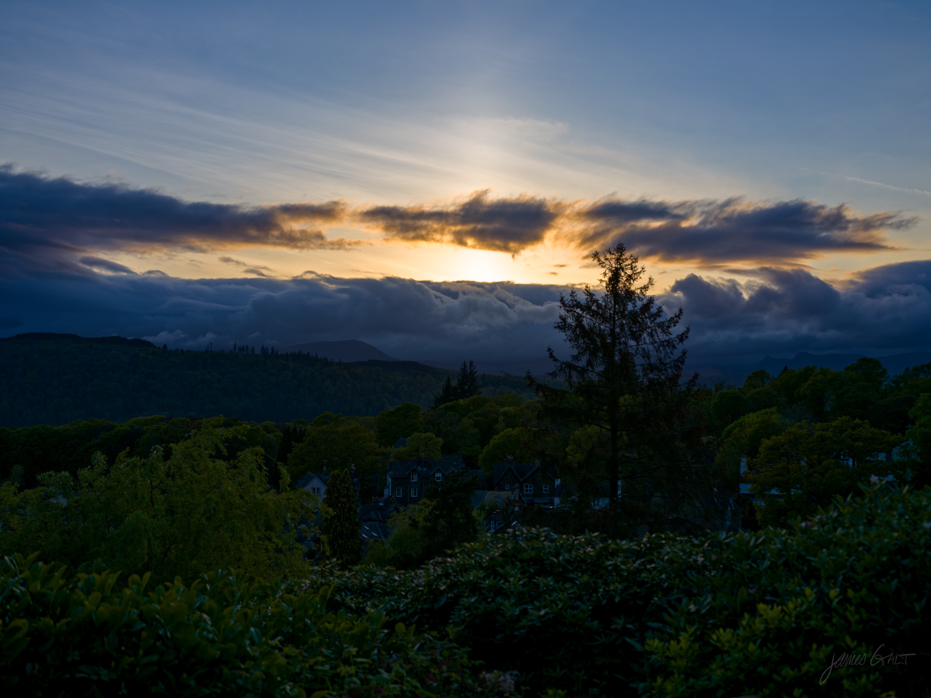 Sunset over the fells seen from a garden, a single conifer silhouetted against gold light breaking under a band of cloud, dark shrubs in the foreground.