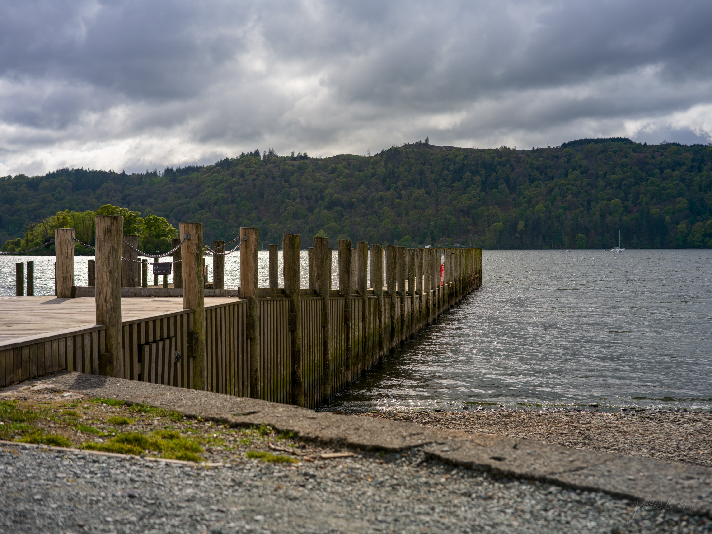 A weathered wooden jetty on thick pilings reaching into a grey lake, dark wooded fellside and low cloud beyond.
