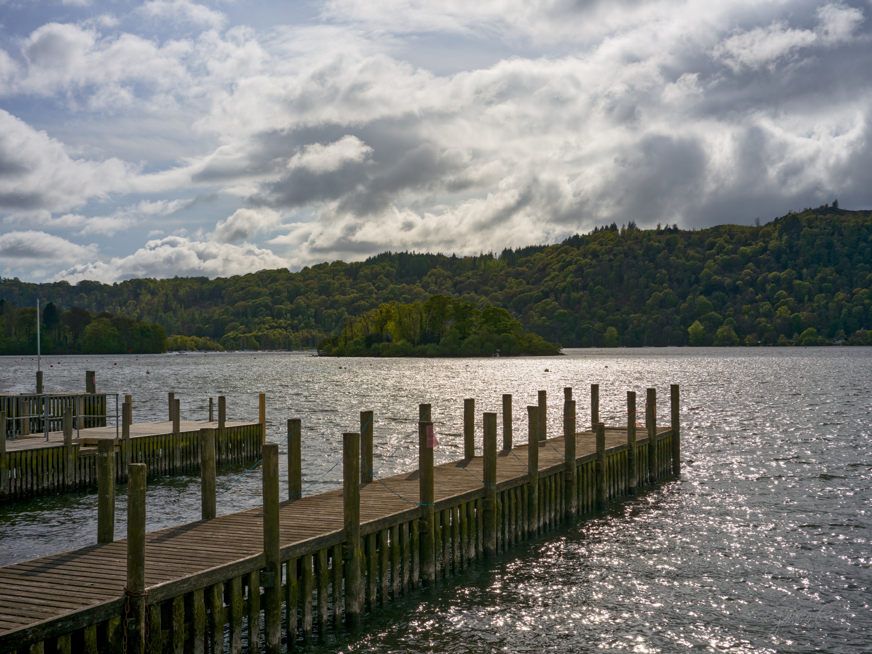 A long wooden jetty running out into Windermere, sunlight breaking through cloud and glittering on the water, a low wooded island in the distance.